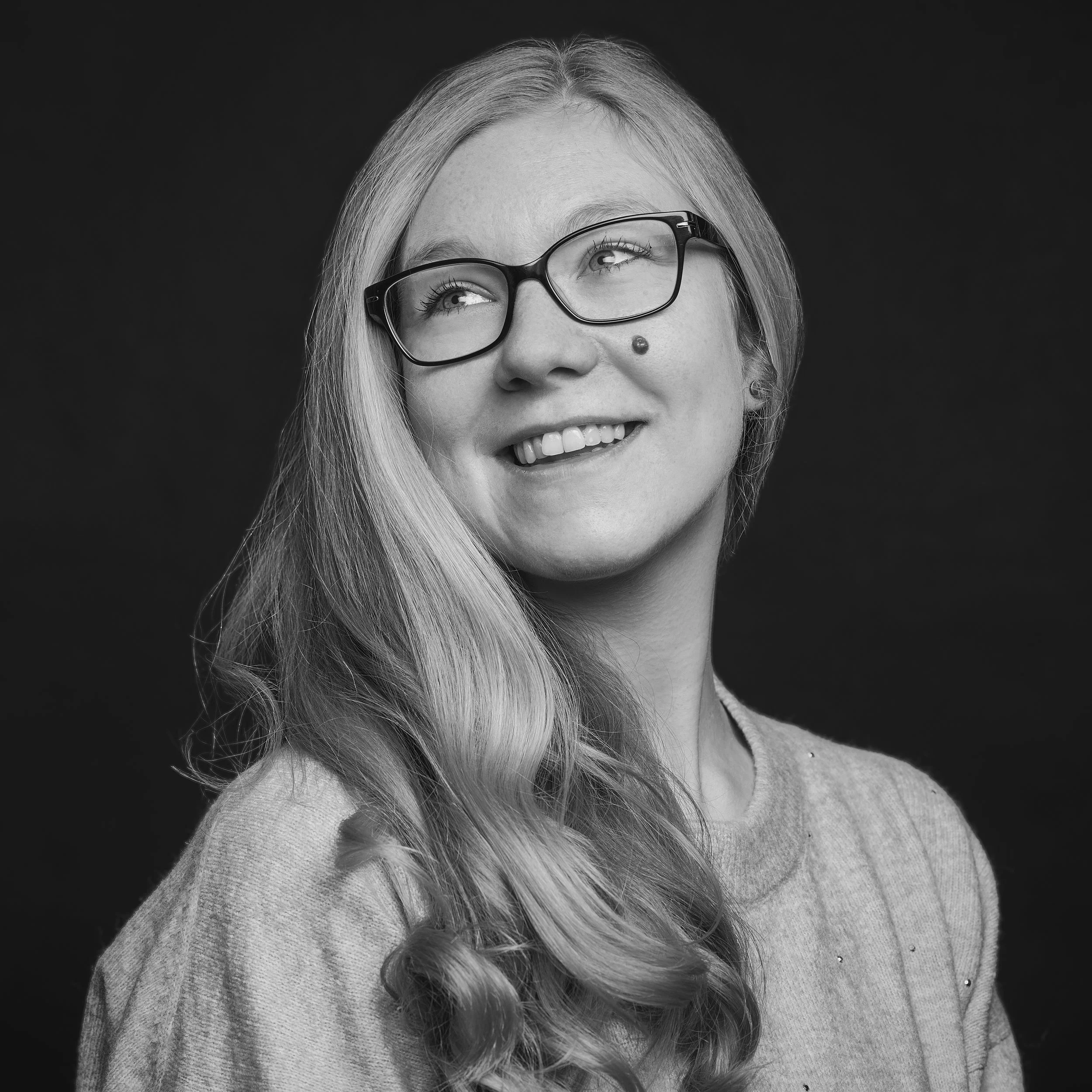 Portrait en noir et blanc d'une femme avec des lunettes, longs cheveux lisses, souriante, portant un pull, avec un fond noir.