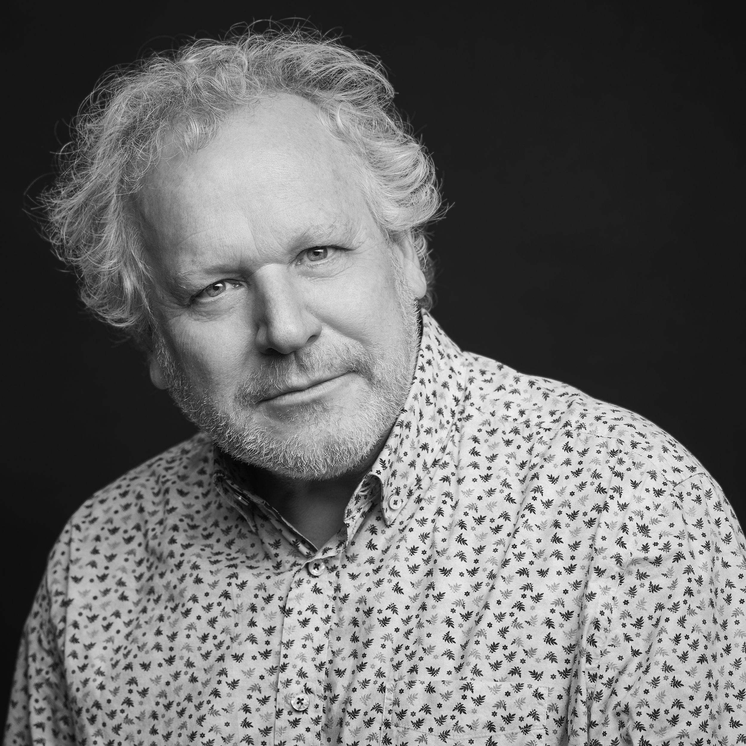 Un homme âgé avec des cheveux gris bouclés et une barbe, portant une chemise à motif floral, regarde directement l'objectif dans une photo en noir et blanc.