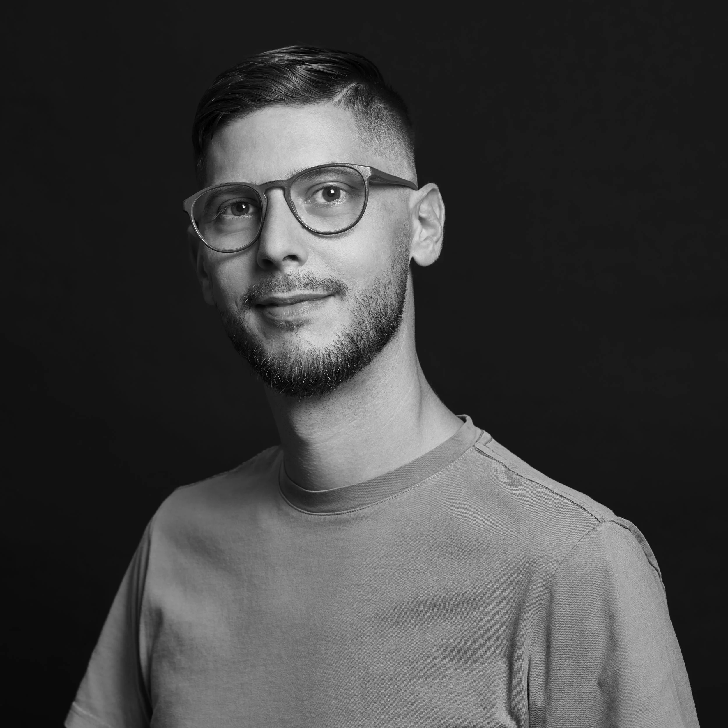 Portrait d'un homme avec des lunettes, barbe, cheveux courts coiffés en arrière, portant un t-shirt gris, fond noir.