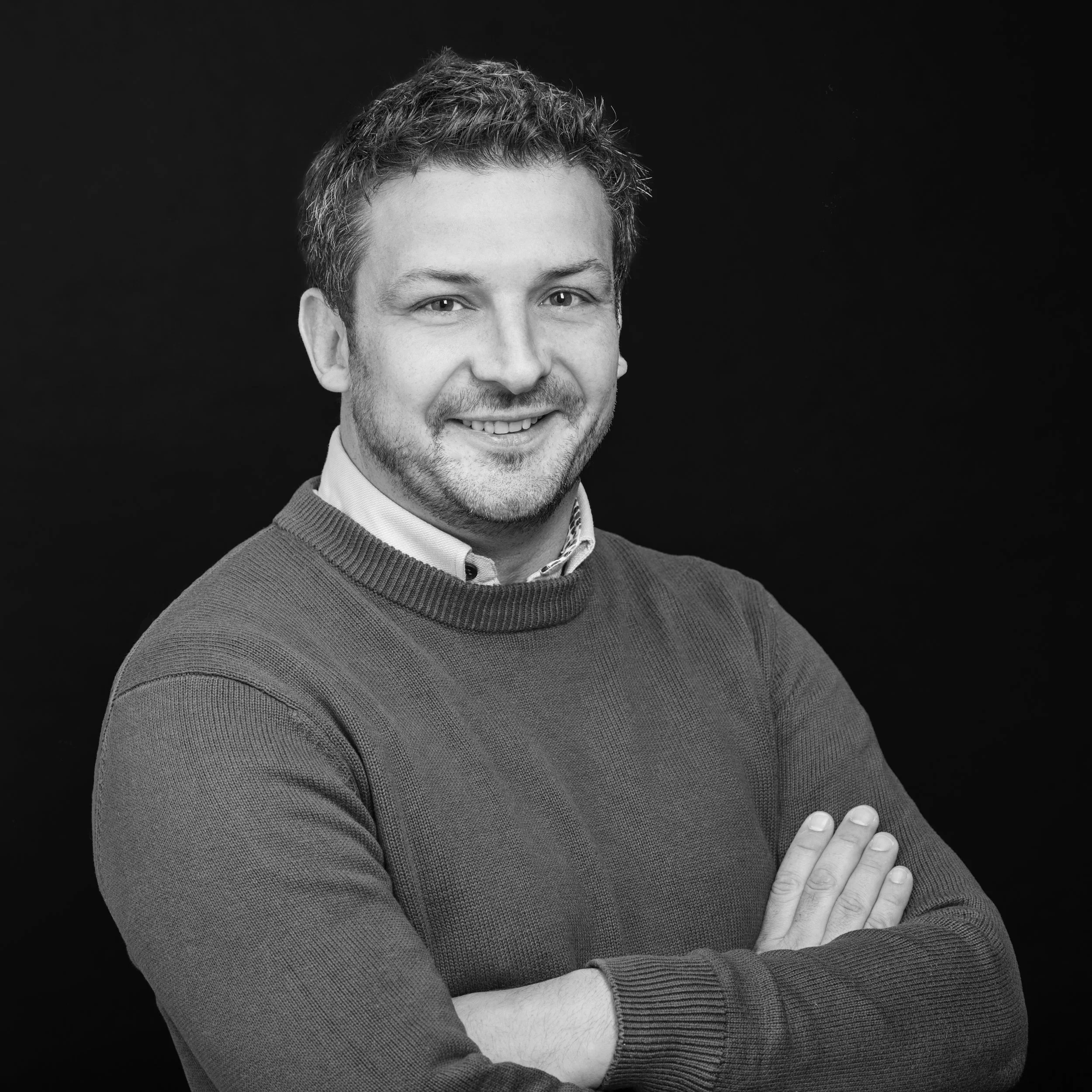 Portrait en noir et blanc d'un homme souriant avec des cheveux bouclés, portant un col roulé et un sweater, croisé les bras, contre un fond noir.