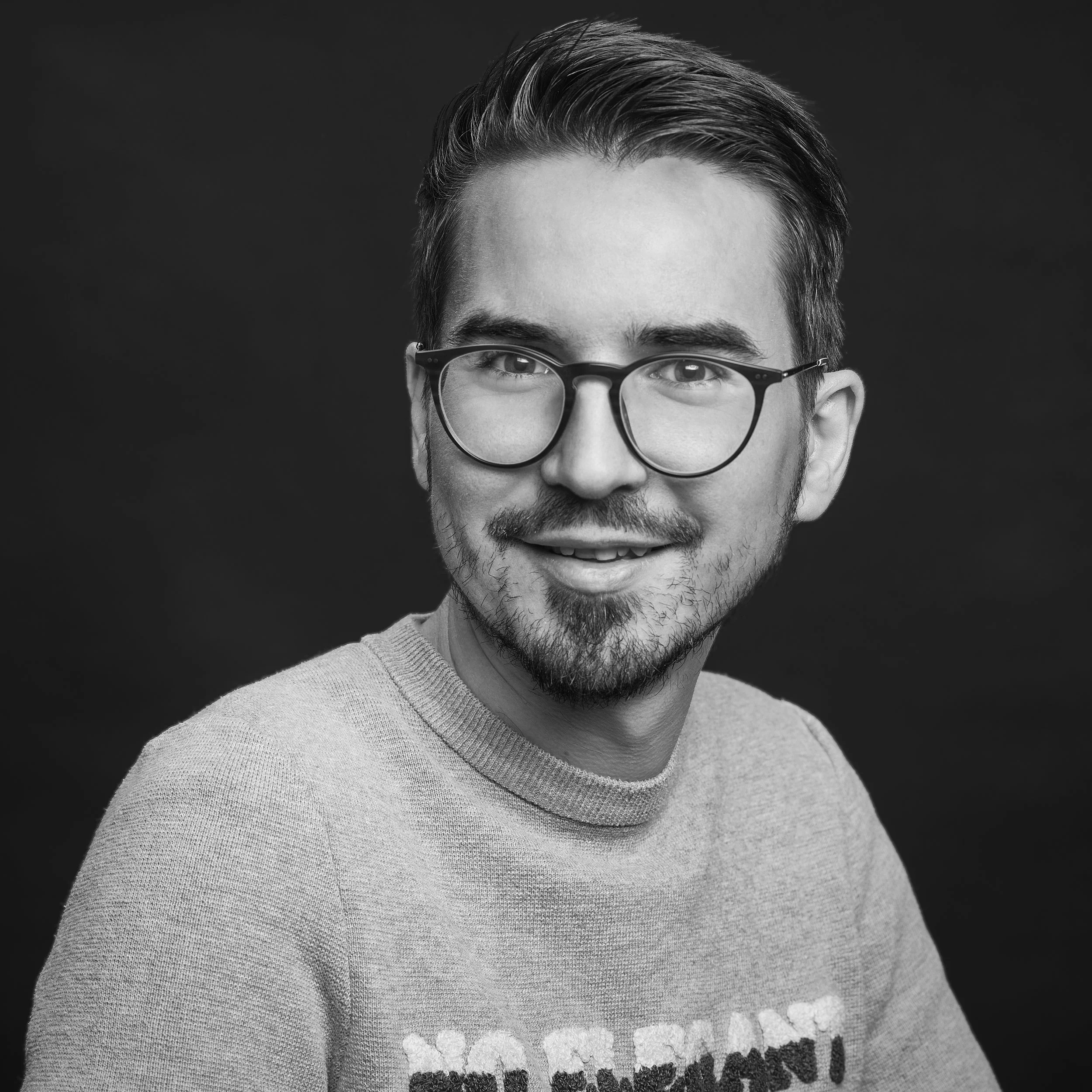 Un portrait en noir et blanc d’un homme avec des cheveux courts et une barbe, portant des lunettes rondes, souriant légèrement, avec un pull gris clair.