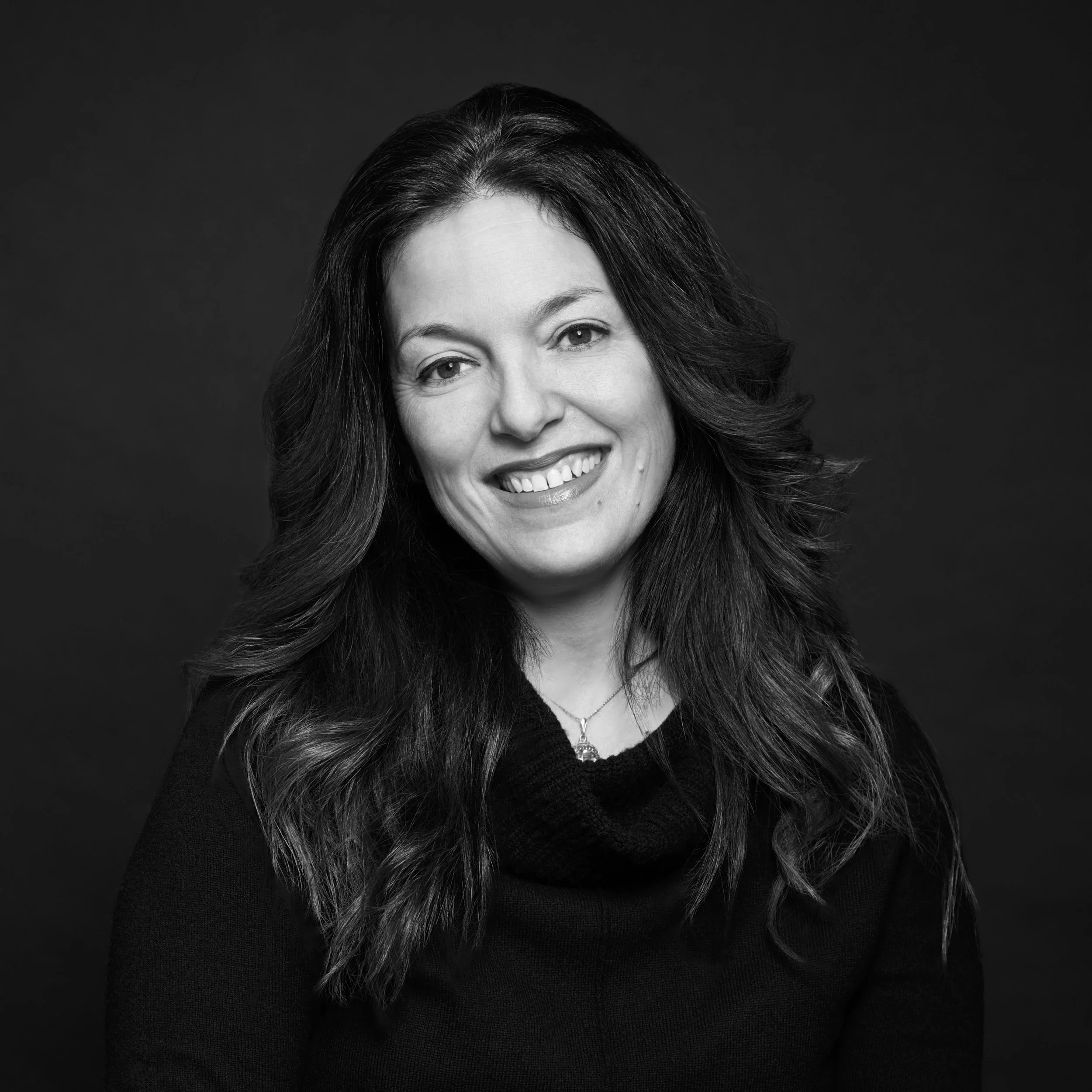 Portrait en noir et blanc d'une femme avec des cheveux longs et bouclés, portant un collier, souriant légèrement. Fond sombre.