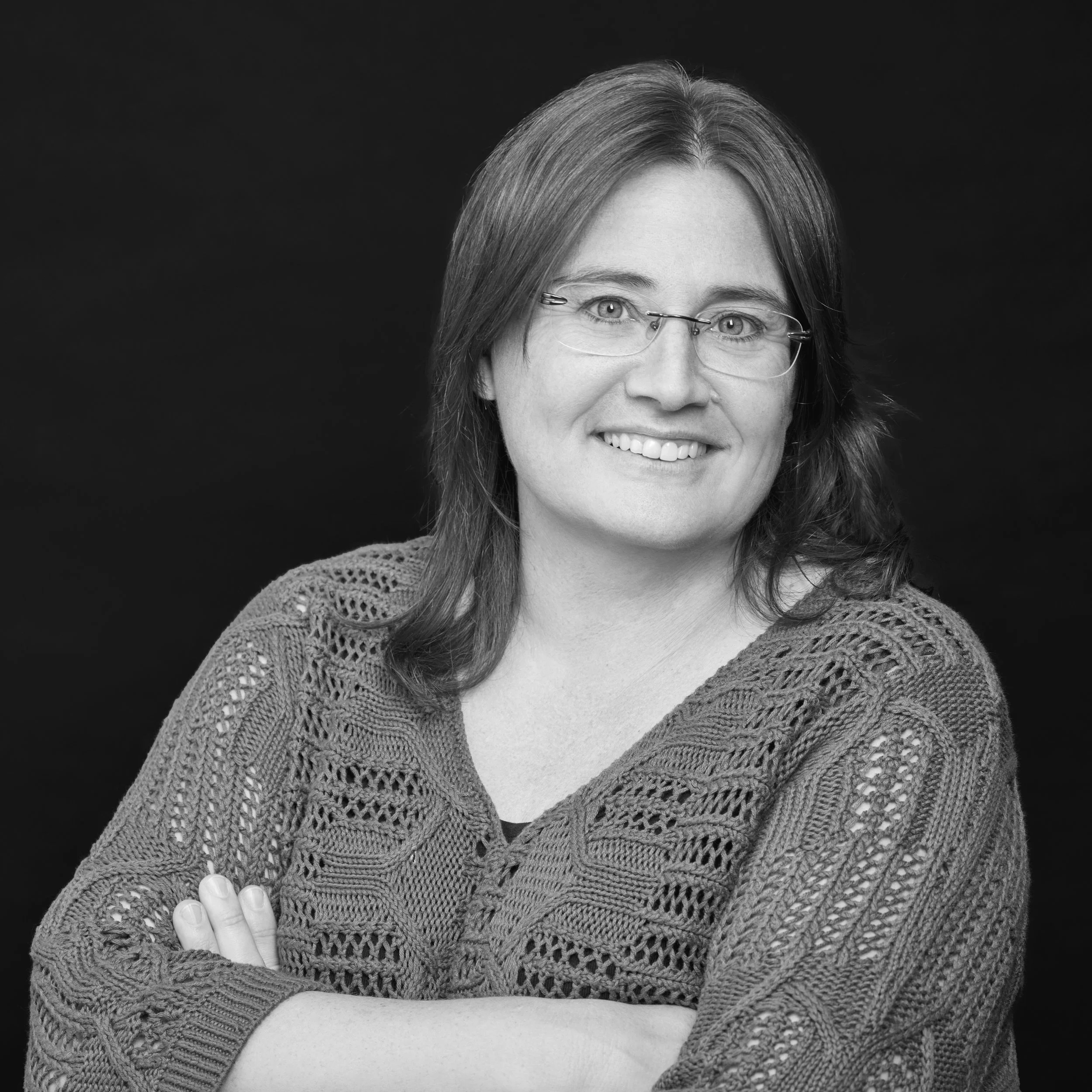 Portrait d'une femme souriante avec des cheveux mi-longs, portant des lunettes et un tricot en crochet, contre un fond noir.