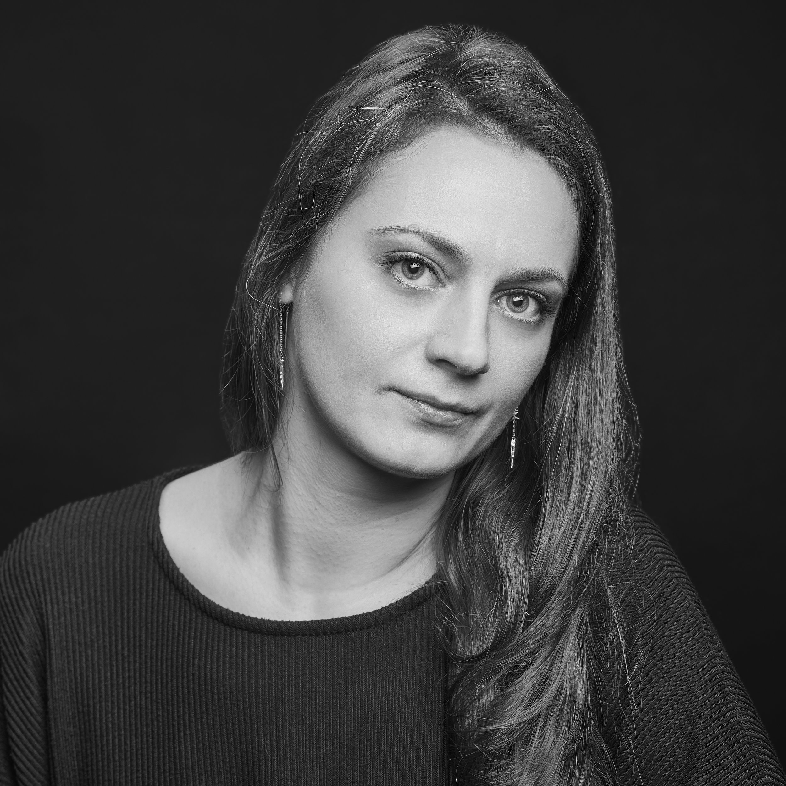 Un portrait en noir et blanc d'une femme avec cheveux longs, portant un pull côtelé, regardant vers l'objectif, fond sombre.
