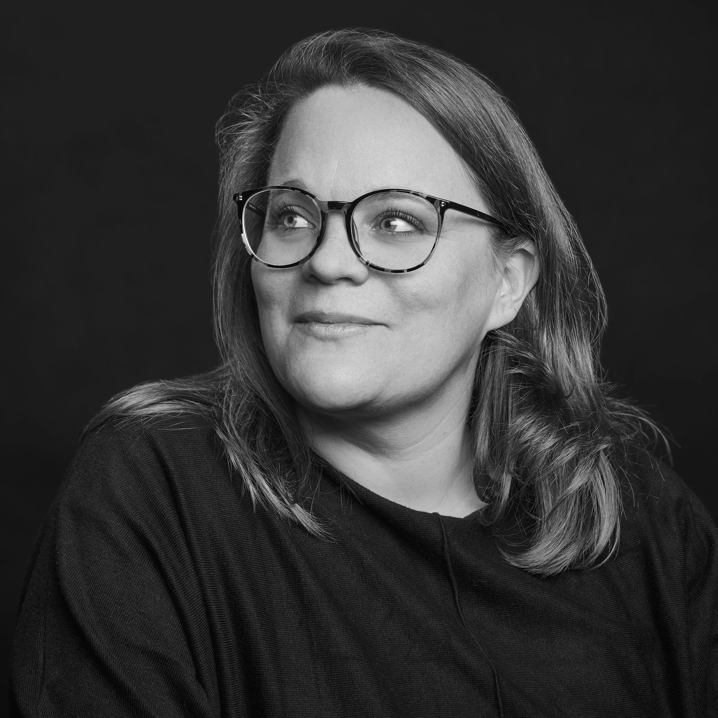 Jeune femme avec des cheveux longs et ondulés portant des lunettes à monture écaille, regardant légèrement vers la droite sur un fond noir et blanc.