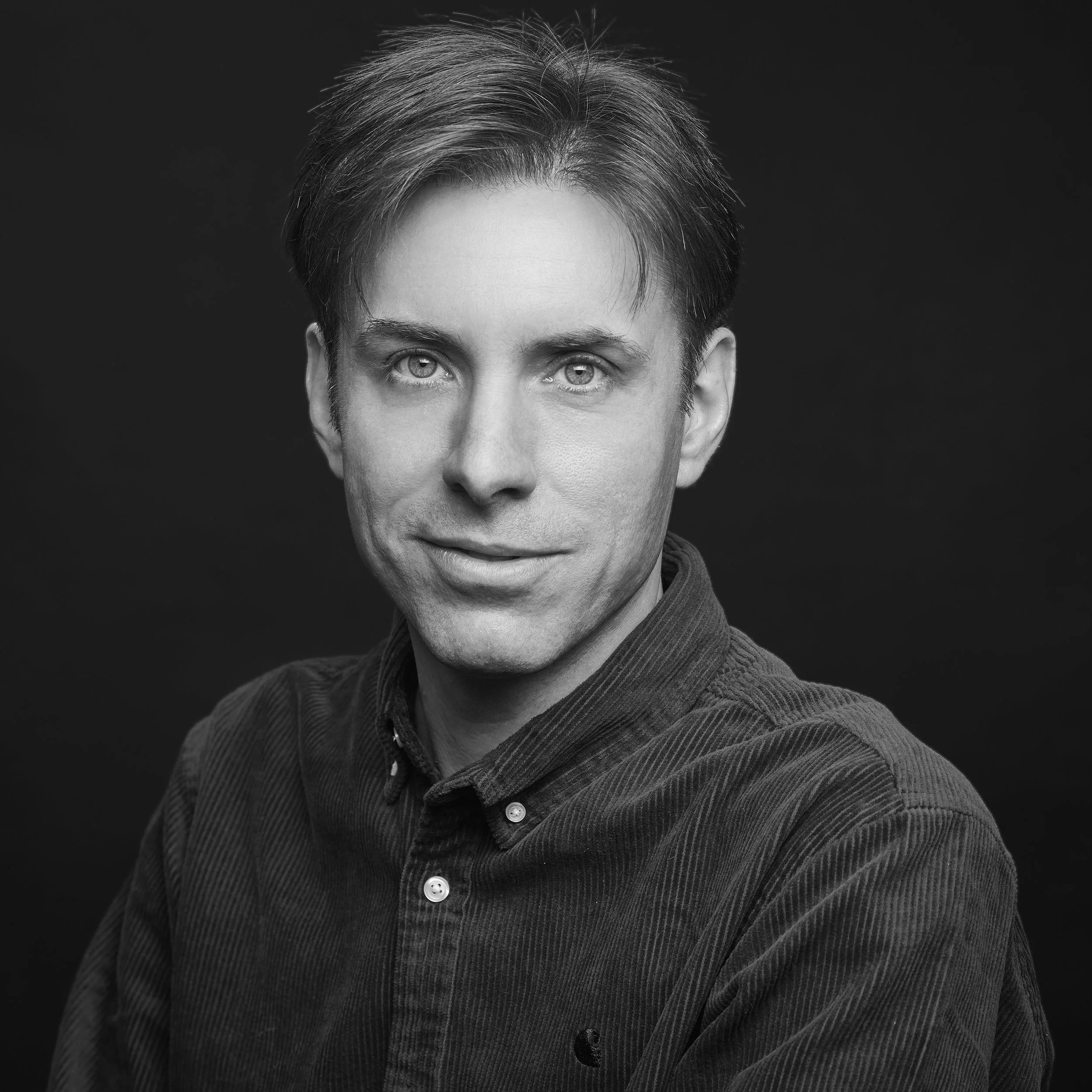 Un portrait en noir et blanc d'un homme avec des cheveux courts et gris, portant une chemise à col et un pull à rayures, contre un fond noir.