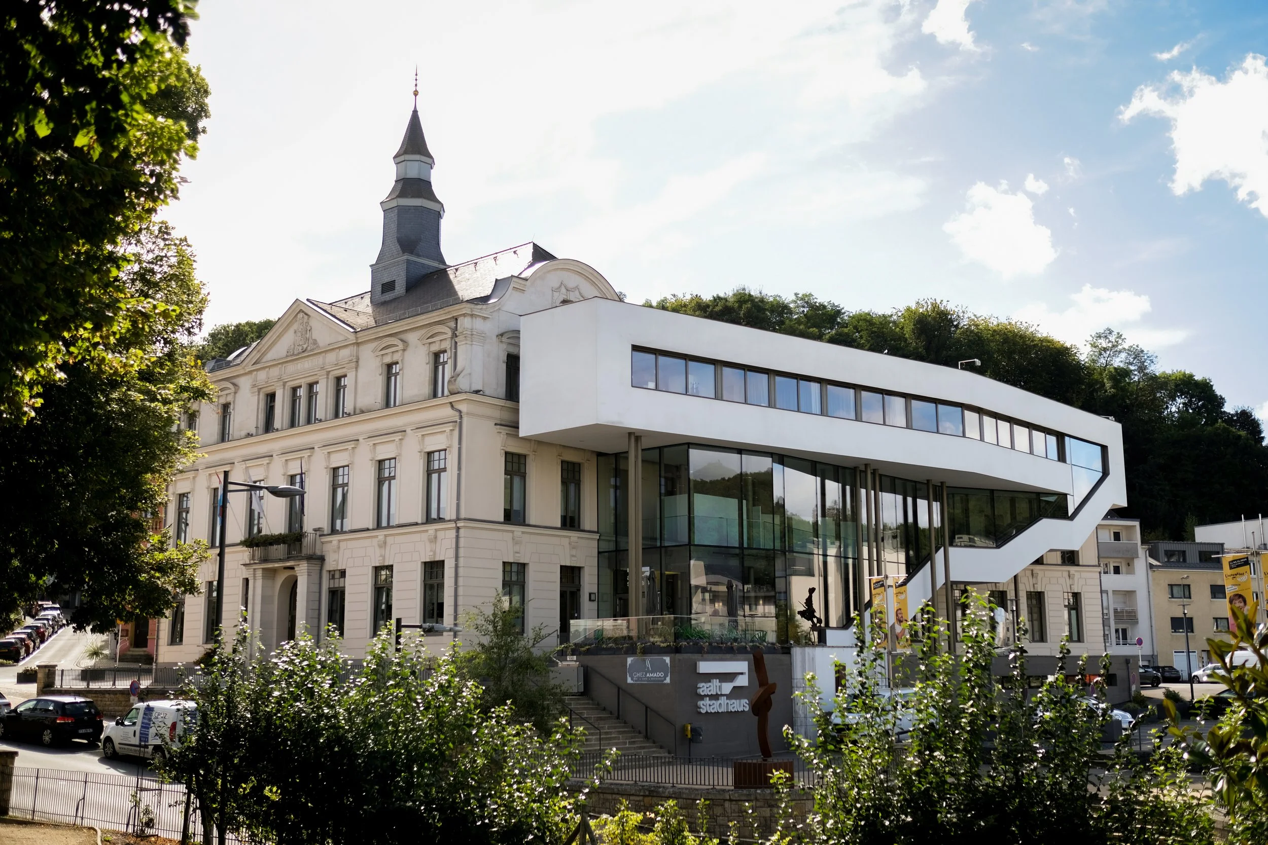 Un bâtiment moderne et un ancien avec une église en arrière-plan, sous un ciel en partie nuageux.
