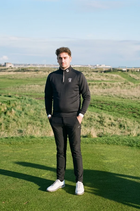 Young man standing on a golf course wearing black athletic jacket and pants, white shoes, with a background of green grass, fields, and a blue sky with some clouds.