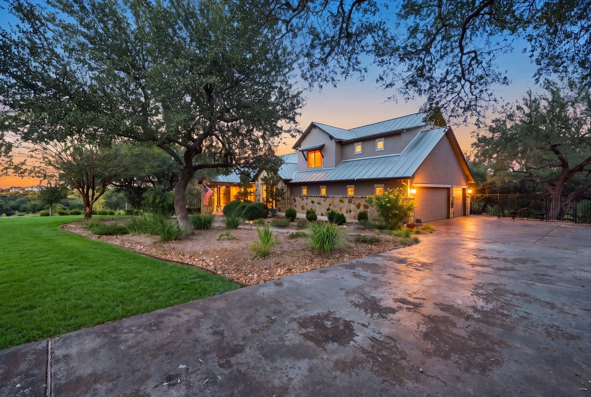 Two story Hill Country home with stone and a metal roof under oak trees at twilight