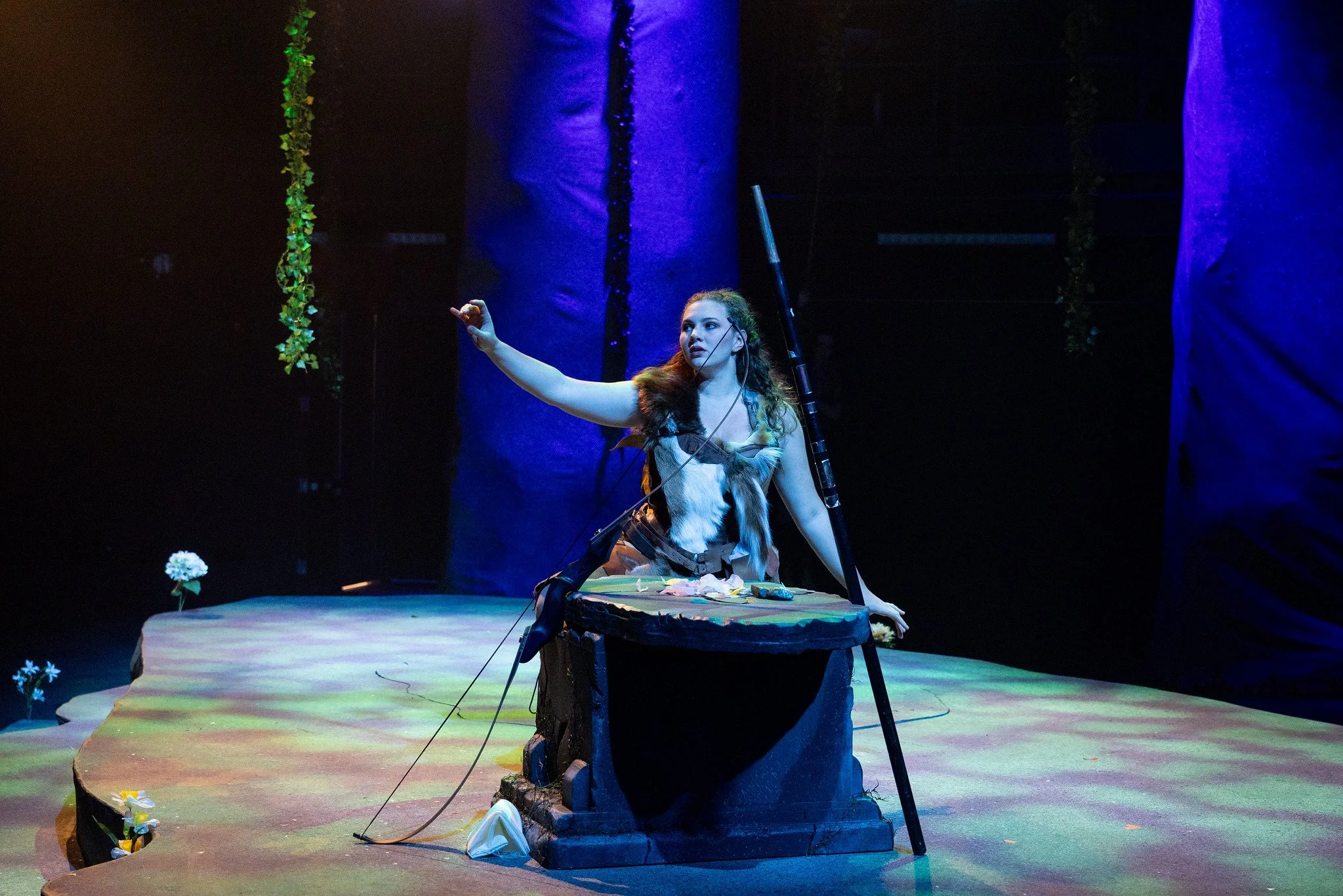 A woman with long dark curly hair and light skin, dressed in a costume resembling a forest creature, is on stage with her arm extended. She is surrounded by a set that includes trees and flowers, lit with colorful stage lighting, creating a mystical atmosphere.