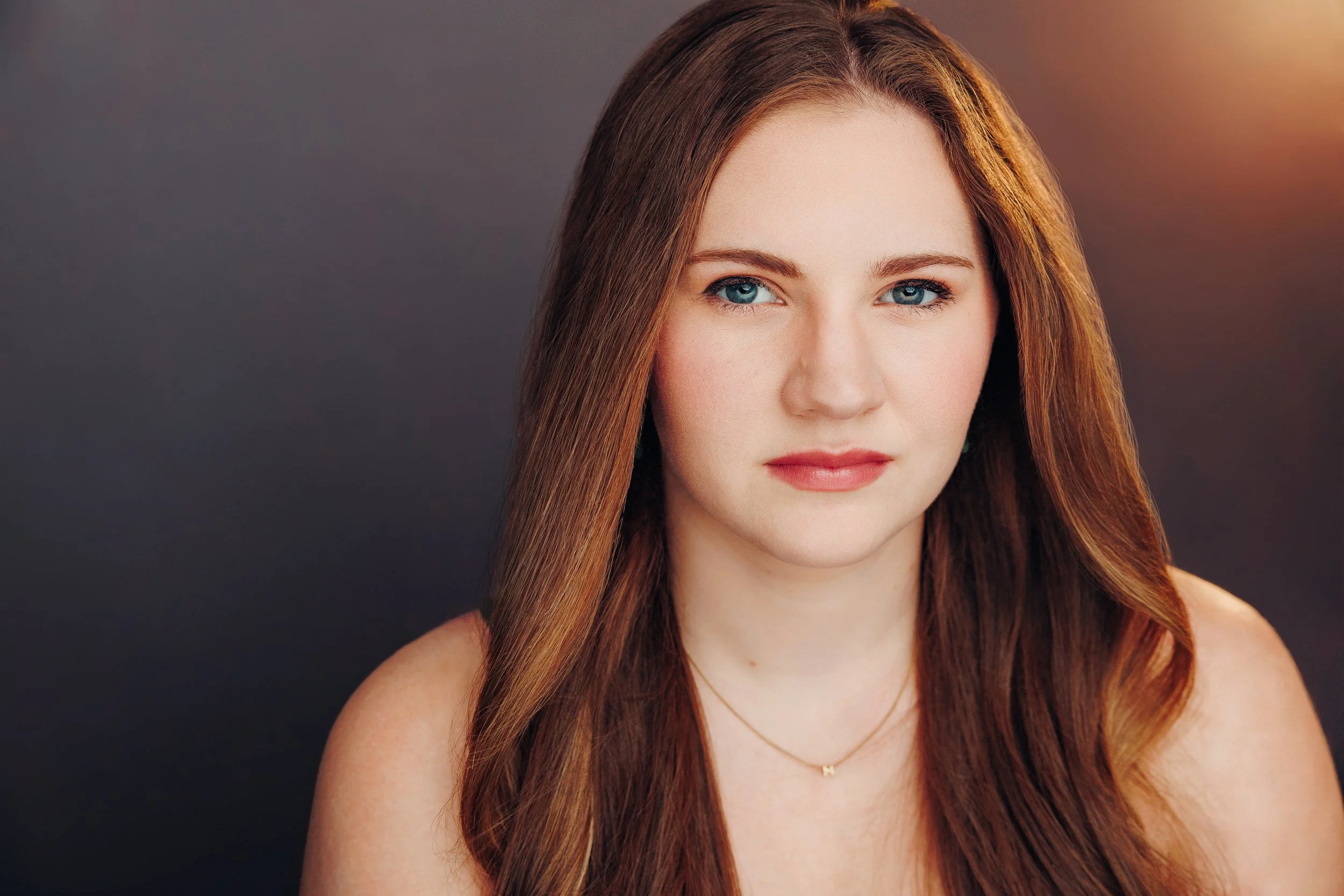 Close-up portrait of a young woman with long red hair and blue eyes, wearing a small gold necklace, against a dark background with a reddish glow on the right.