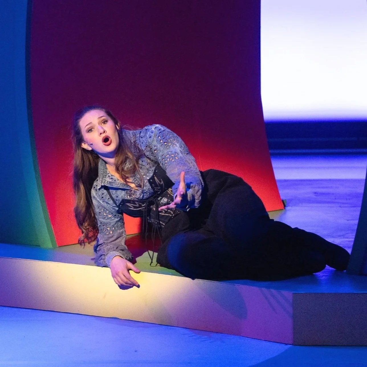 A woman performing on stage, sitting on the floor with her body turned to the side, wearing a patterned blouse and black pants, with her hair down, expressive face, and gesturing with one hand.
