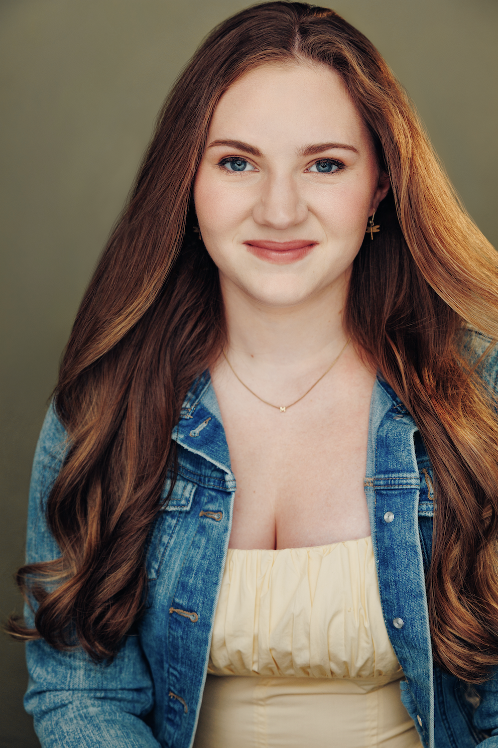 A young woman with long red hair, blue eyes, and light skin, smiling softly, wearing a yellow top, denim jacket, a necklace with an 'M', and a small earring, against a plain green background.