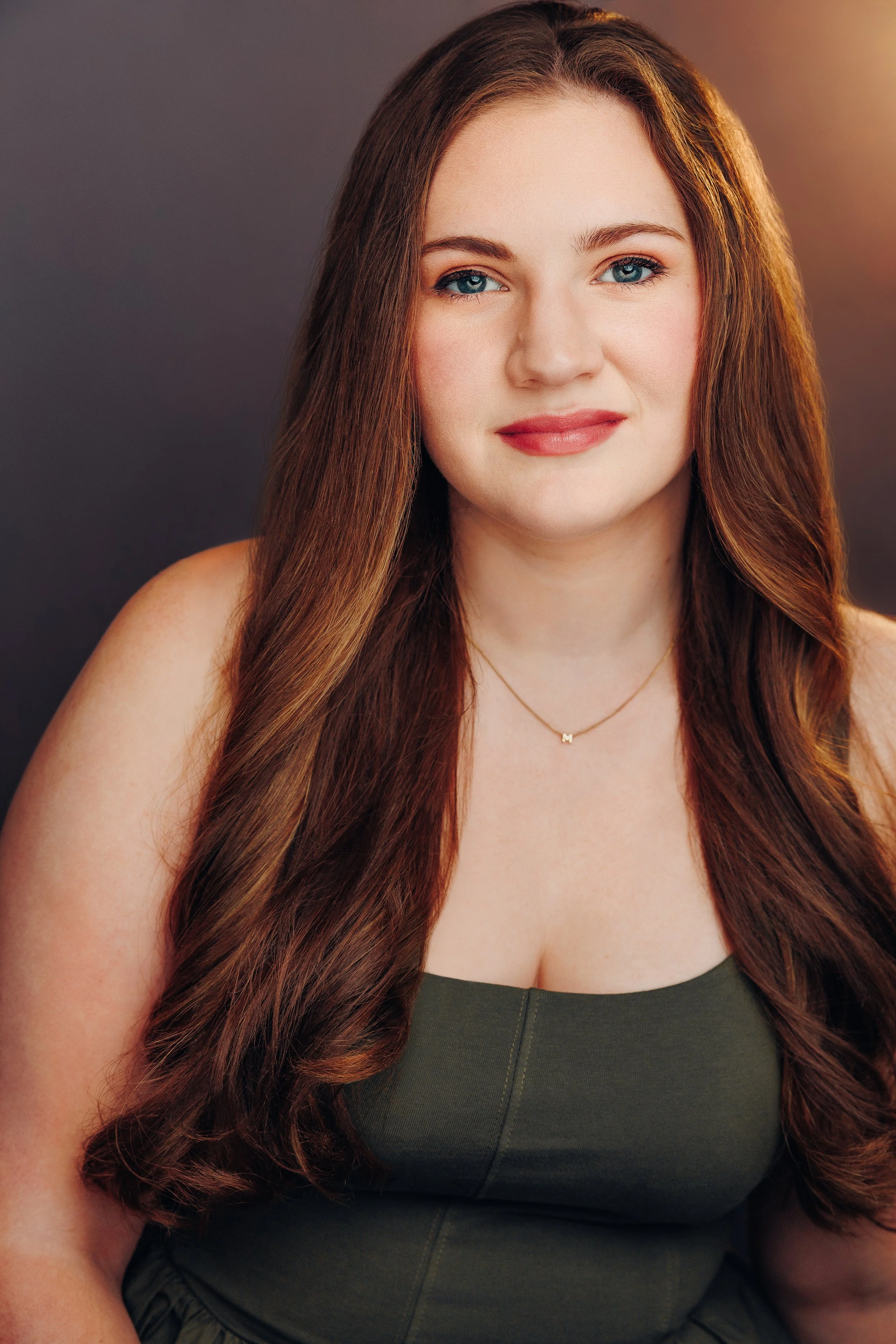 A young woman with long, wavy red hair and blue eyes, wearing a dark green strapless top and a delicate gold necklace with a small letter 'M', smiling softly with a neutral background.
