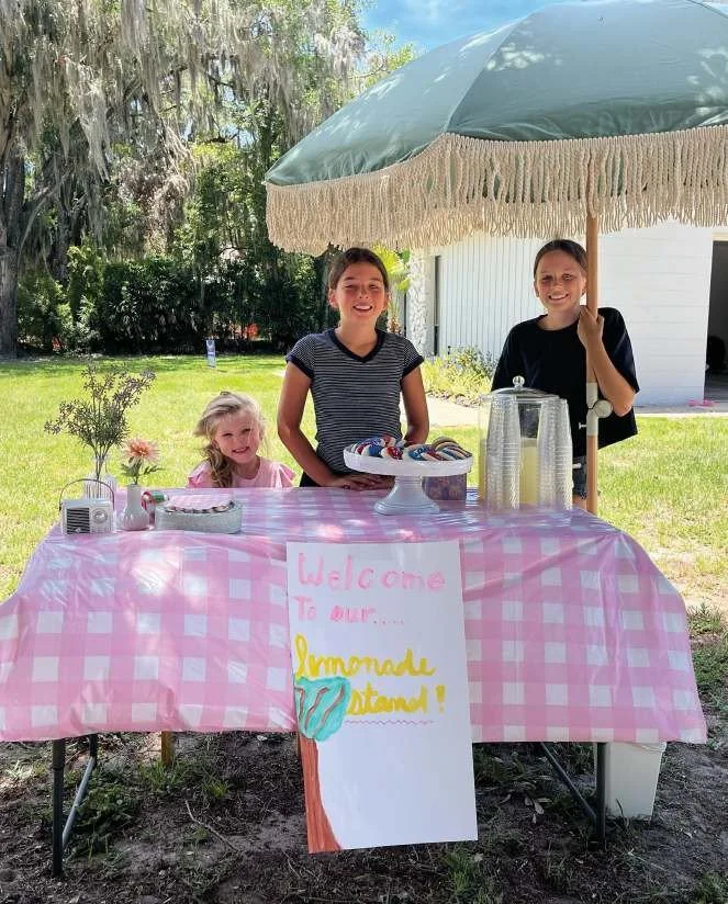 Three young girls at their lemonade stand under a sage-green fringed umbrella in Windermere