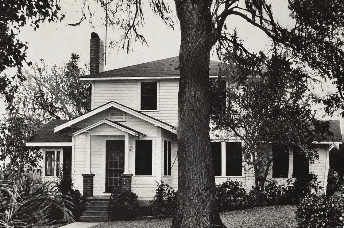 Black-and-white photo of a two-story wood-frame house beside a large tree