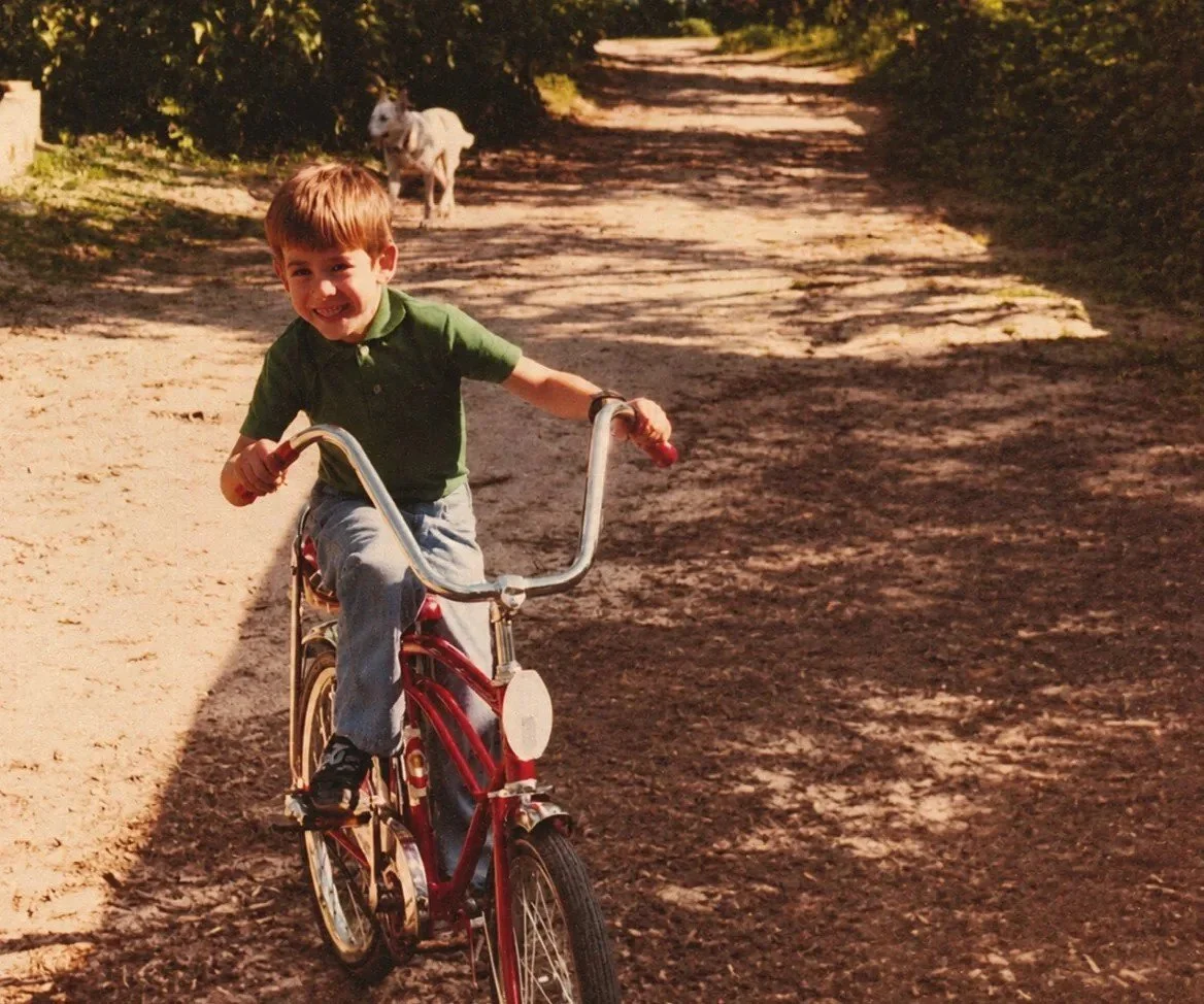 Vintage color photo of a young boy riding a red bicycle down a sandy dirt road under trees