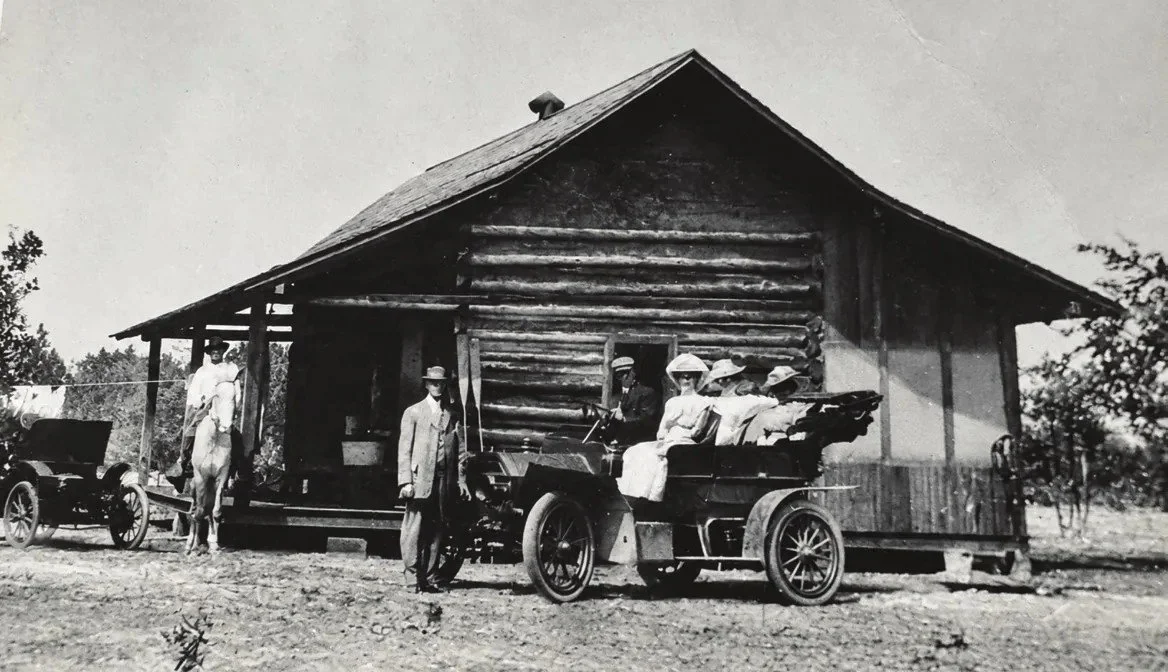 Early 1900s black-and-white photo of a log house with a wide porch, with people in period dress and two antique automobiles out front