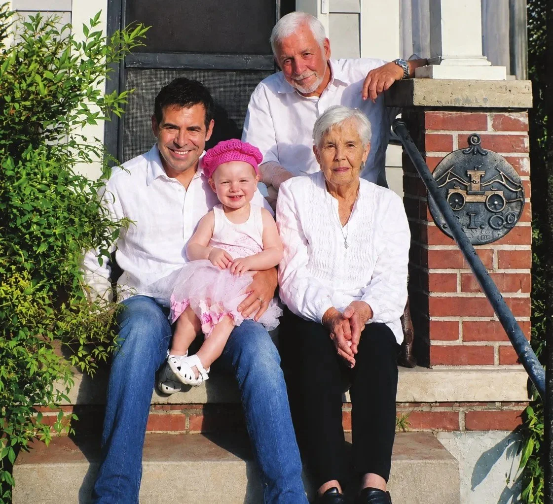 Four generations of a family seated on the brick front steps of a house: an older couple, a younger man, and a toddler girl in a pink dress