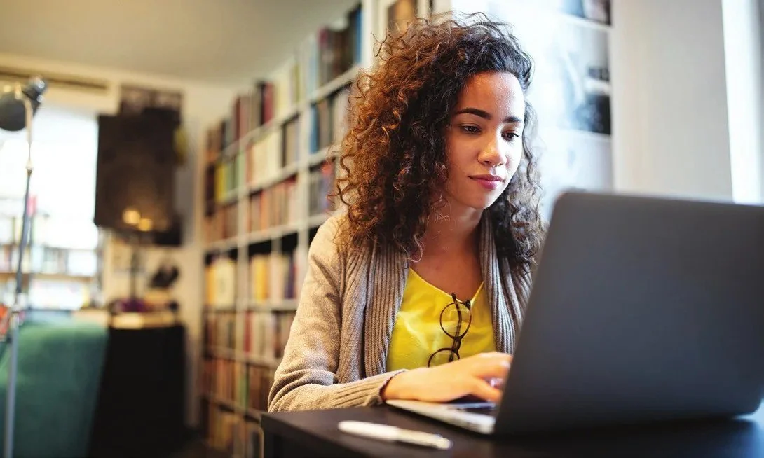 A young woman with curly hair studying on a laptop in front of tall bookshelves