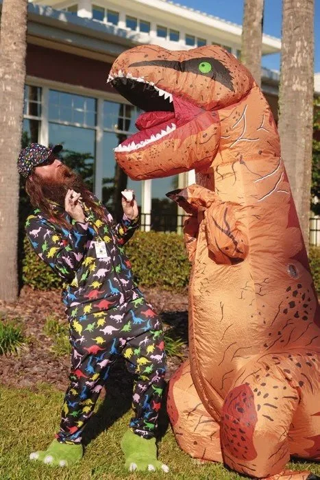 A person in an inflatable T-rex costume roars playfully beside a person in dinosaur-print pajamas outside a library