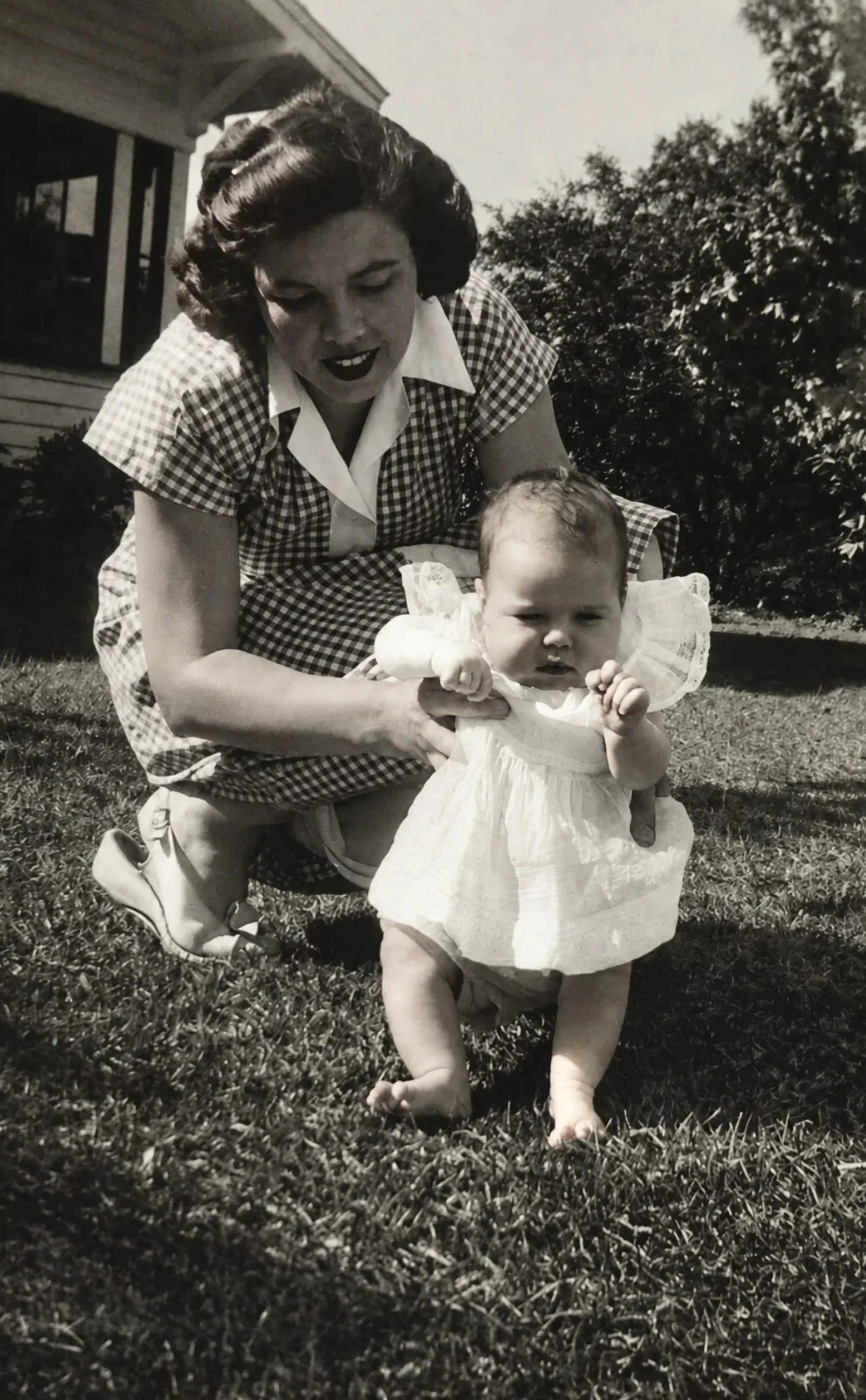Vintage black-and-white photo of a woman kneeling on the grass and steadying a toddler in a white dress who is learning to walk