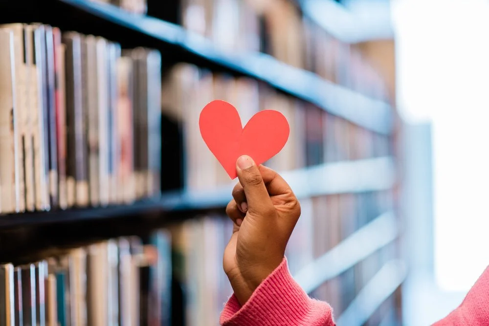 Hand holding a paper heart in front of library shelves for Heart Month at the Orange County Library System