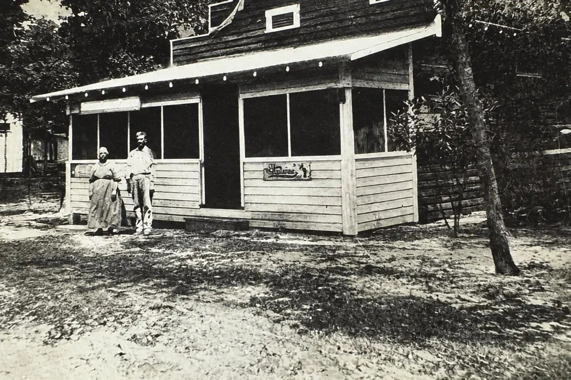 Black-and-white photo of a small wooden building with a screened front porch and two people seated outside