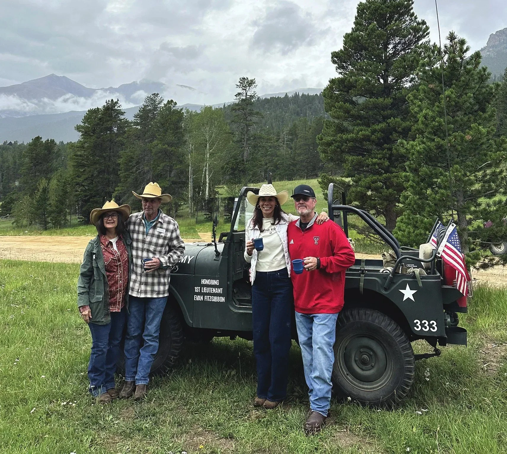 Two couples in western wear standing beside a vintage military jeep with an American flag in a mountain meadow