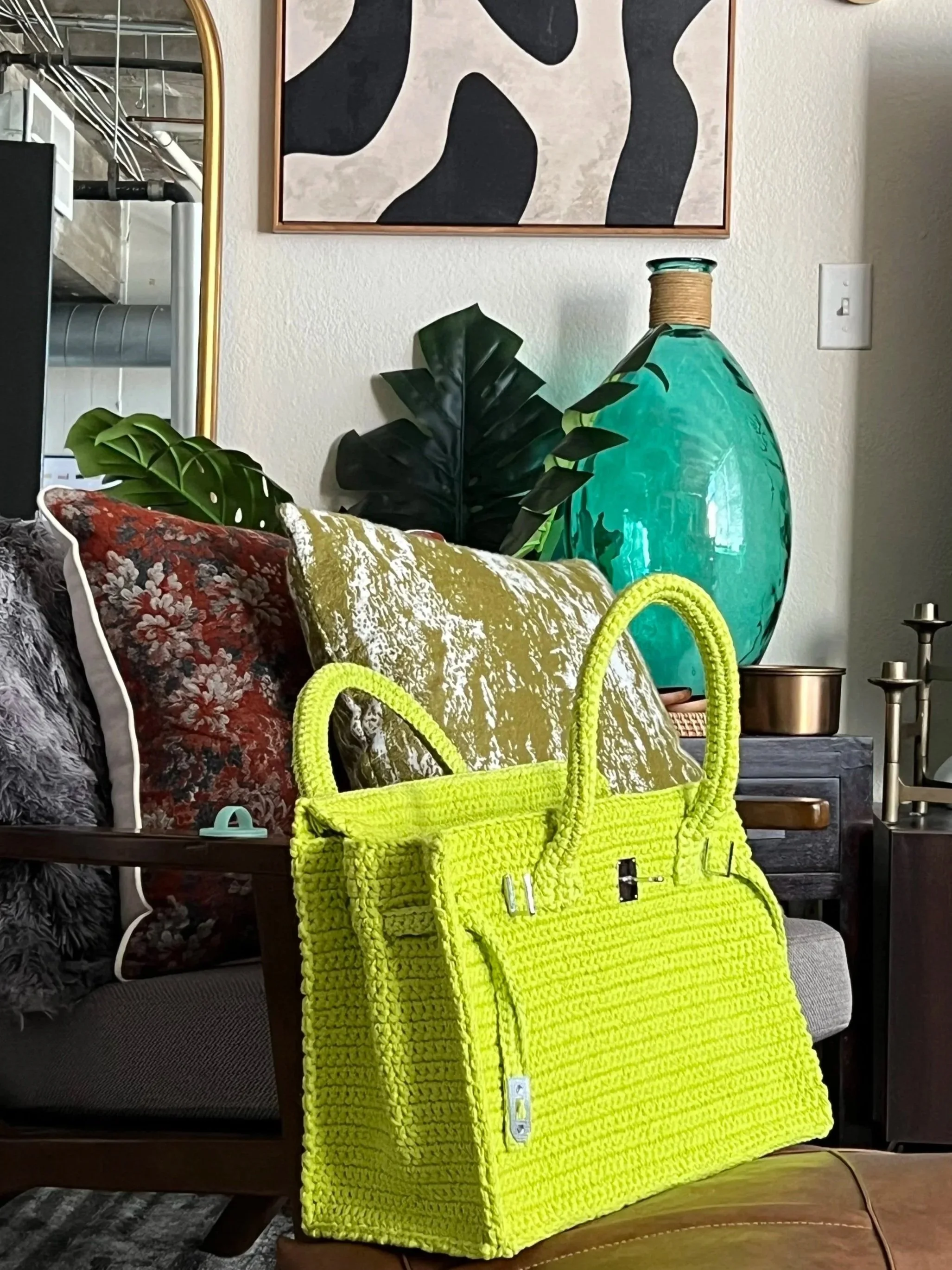 Neon yellow woven handbag displayed on a leather ottoman, surrounded by patterned pillows, tropical leaves, a turquoise glass vase, and framed abstract wall art.