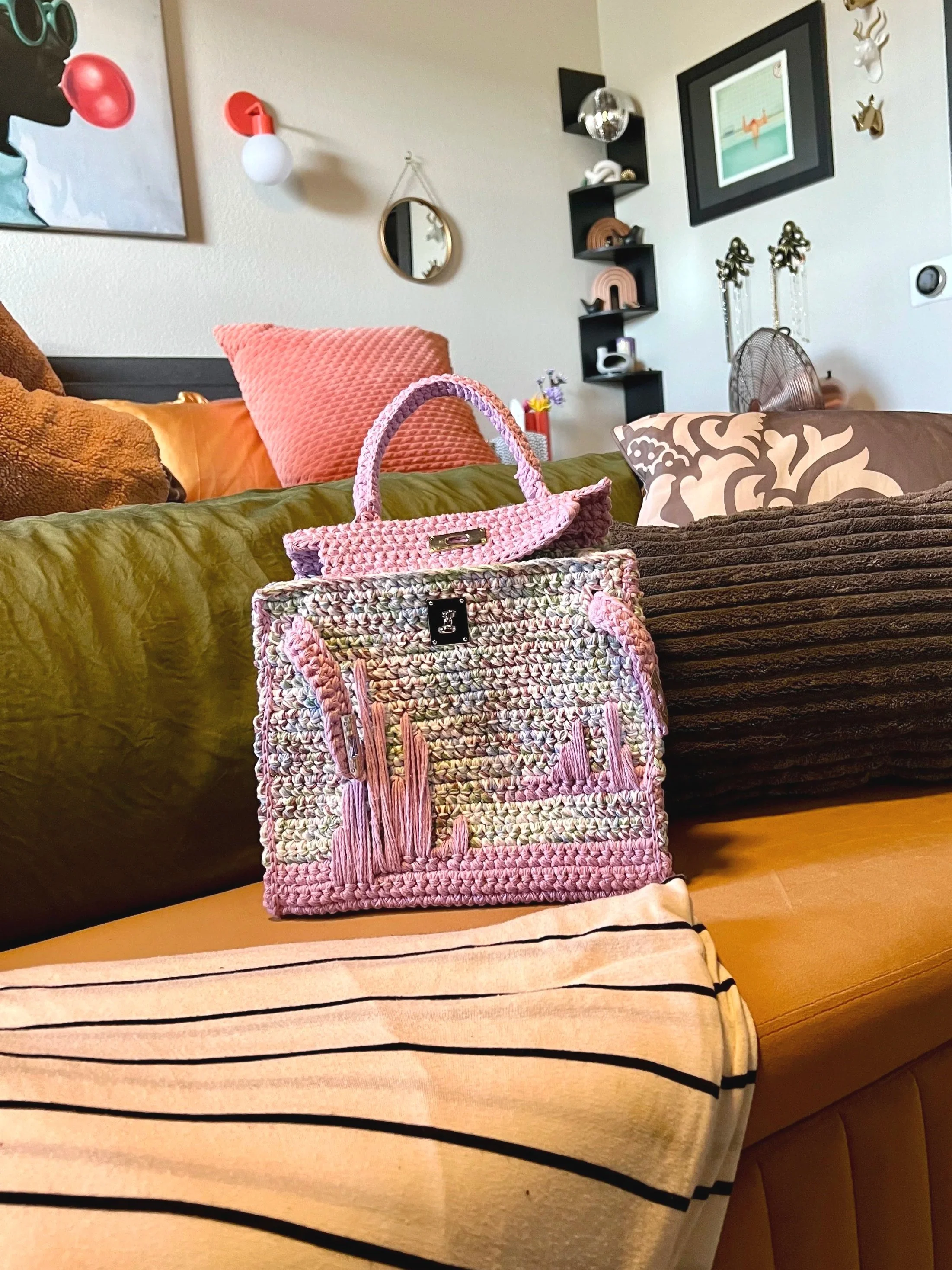 Colorful woven handbag resting on a striped ottoman in a cozy living room with pillows, wall shelves, artwork, and a fan.