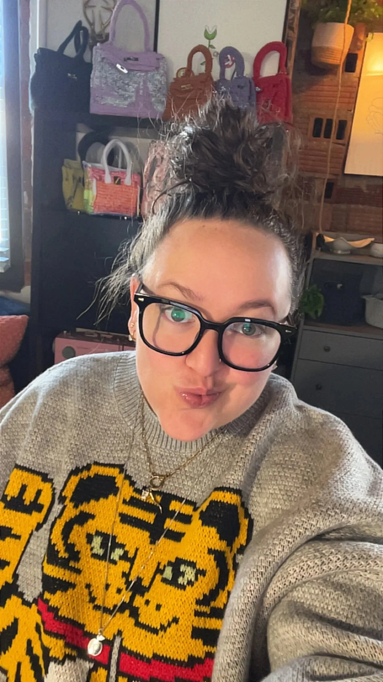 Person wearing black glasses and a gray sweater with a colorful graphic, posing indoors near shelves displaying handbags.