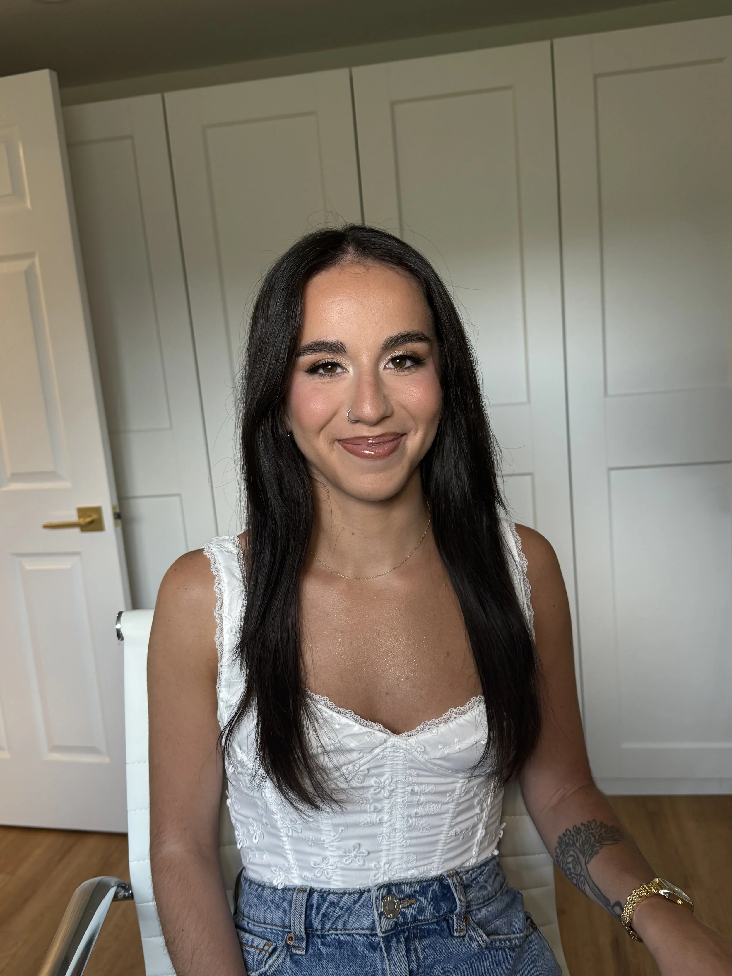 A young woman with long dark hair, wearing a white embroidered top and denim shorts, sitting in a white chair getting her makeup done in front of white cabinets, smiling at the camera, with jewelry including a gold watch, necklace, and nose ring.