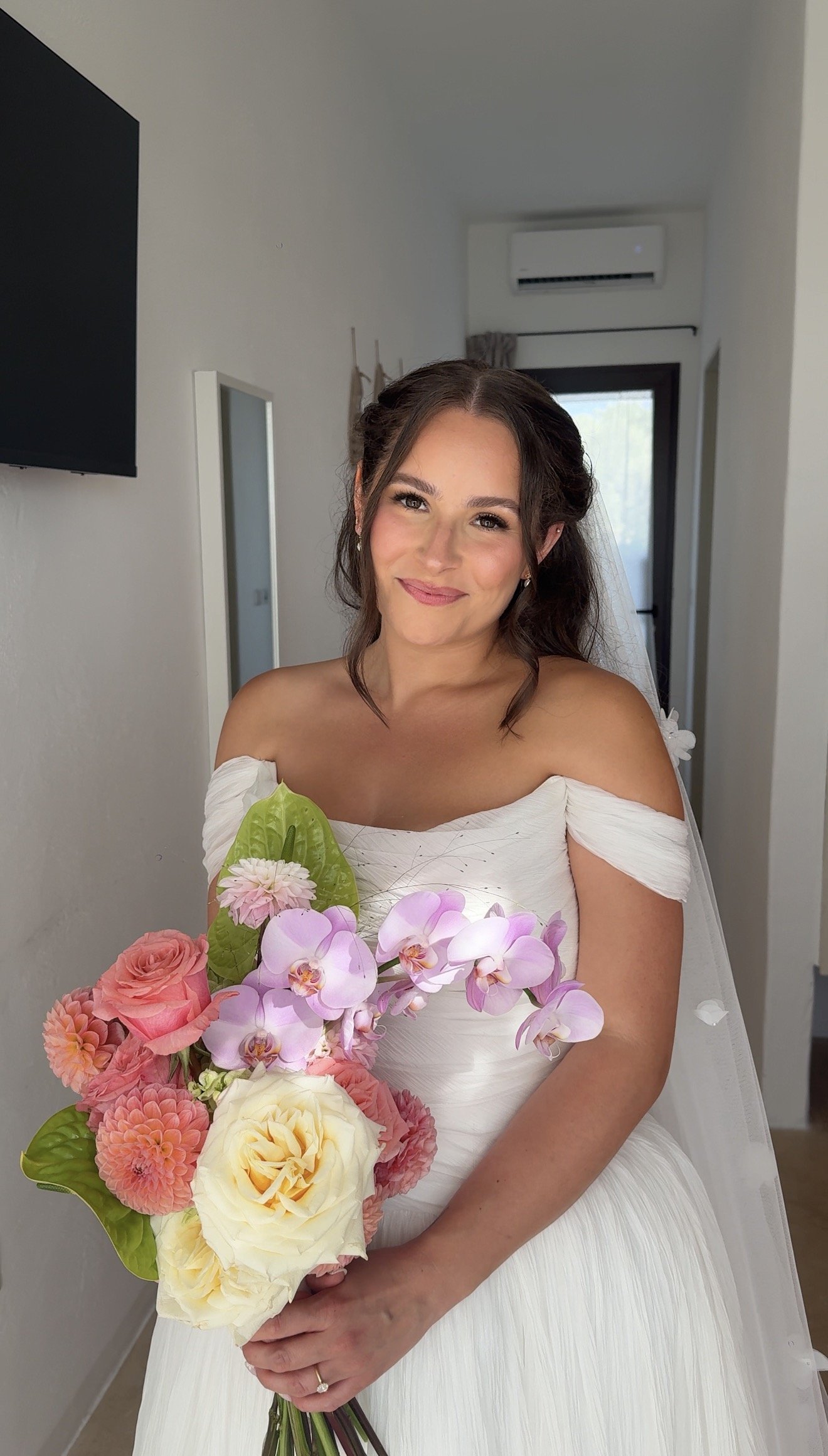 Woman in a white wedding dress smiling and holding a bouquet of pink, purple, and cream flowers with her bridal makeup done.