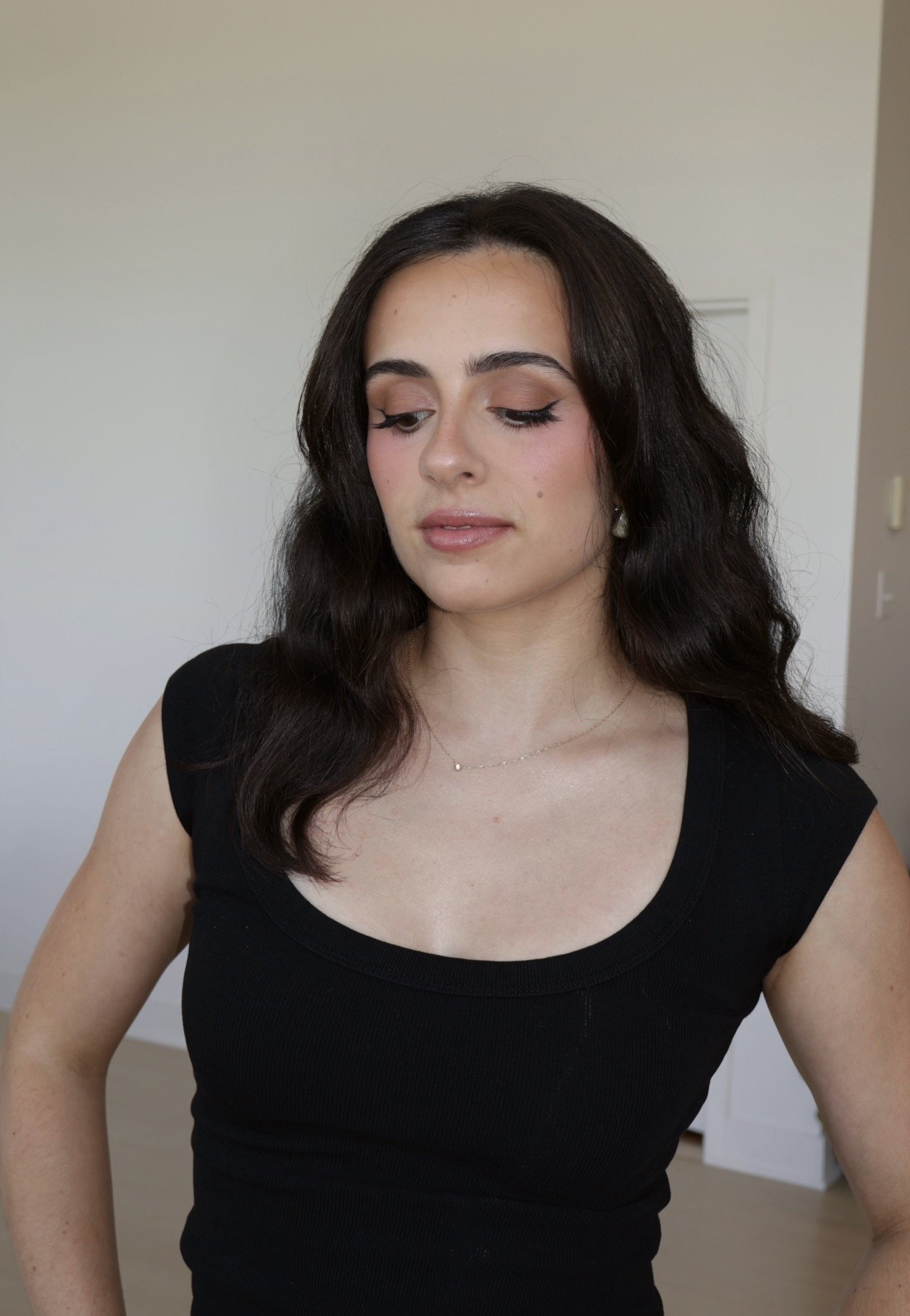 A woman with dark wavy hair, wearing a black top with a scoop neckline, looking down with her eyes closed to show her makeup, standing in a room with plain white walls.
