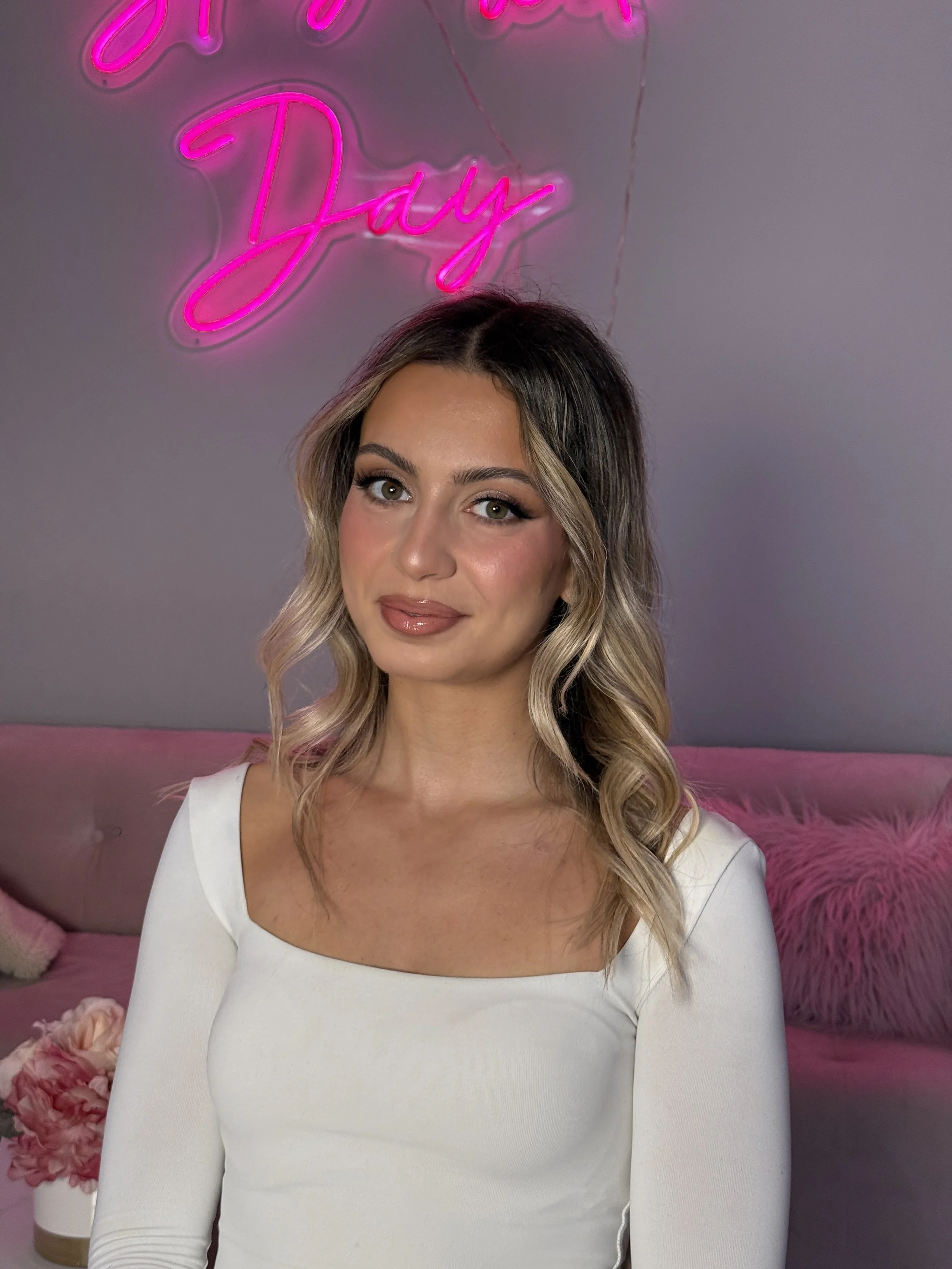 A woman with blonde, wavy hair styled with darker roots, makeup done, wearing a white long-sleeve top, posing indoors with pink floral accents, a pink furry pillow, and a pink neon sign that says 'Slay All Day' in the background.