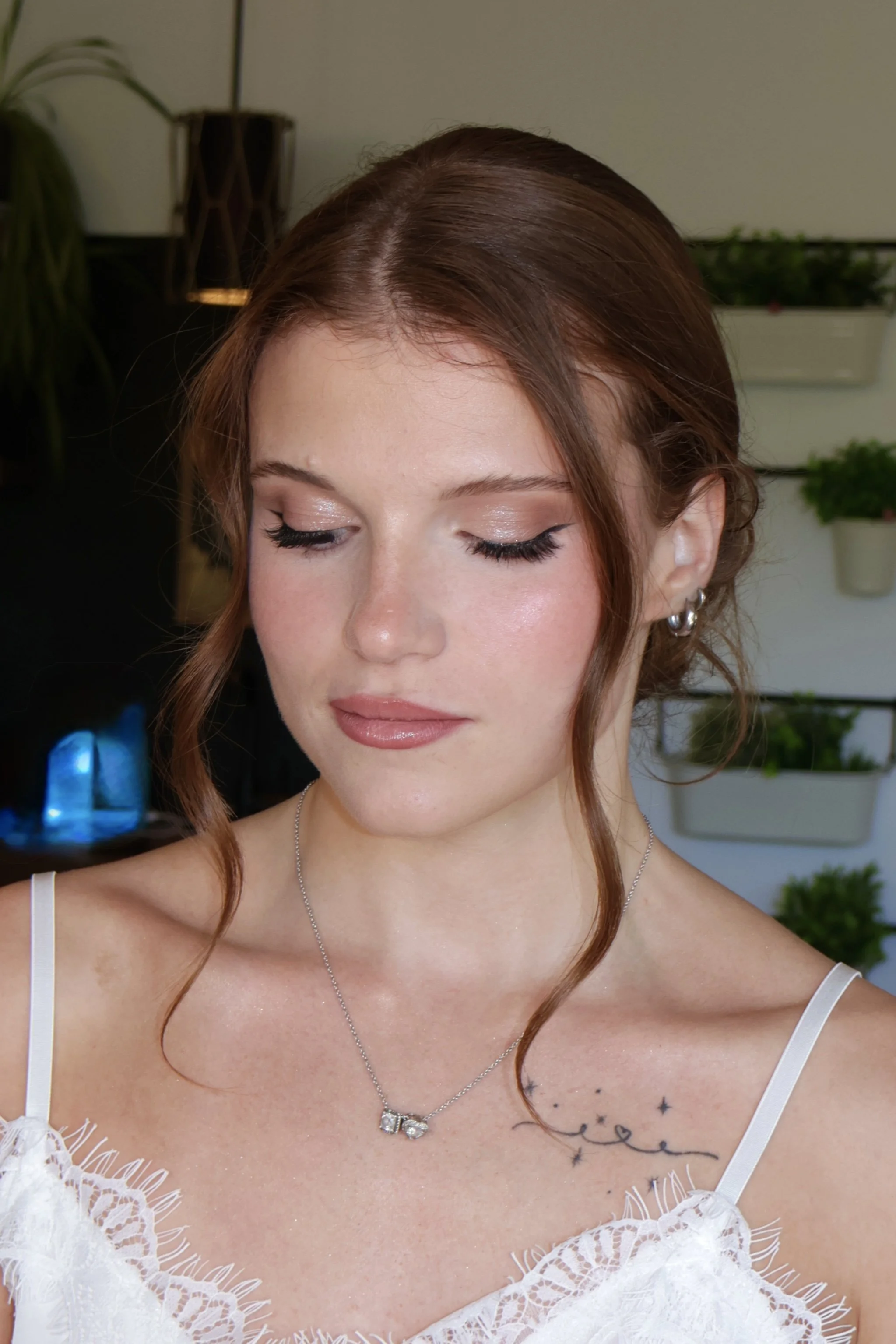 Close-up of a bride with wavy auburn hair, makeup, earrings, a necklace, and a white lace top, with a background of houseplants and a beige wall showing her bridal makeup.
