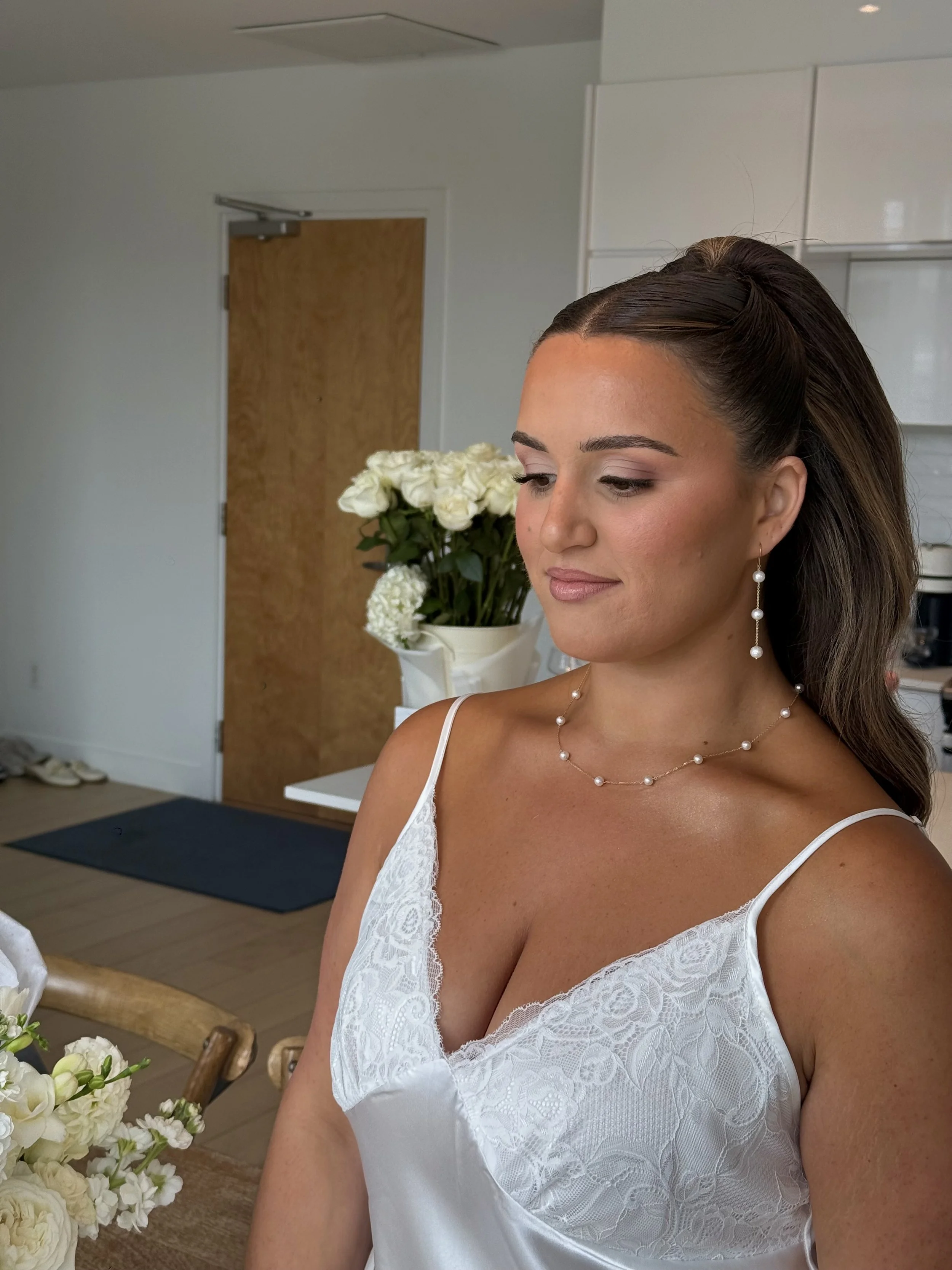 A bride with brown hair styled in an elegant updo, soft pink makeup, wearing pearl jewelry, a white lace PJ, and standing indoors near a table with a bouquet of white flowers. 