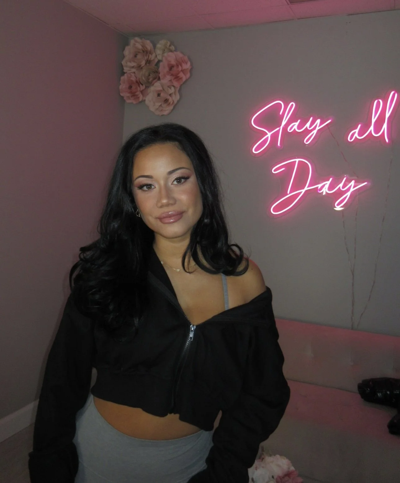 A woman with long dark hair posing to show her makeup in front of a wall with pink neon sign reading 'Slay all Day' and decorative pink paper flowers.