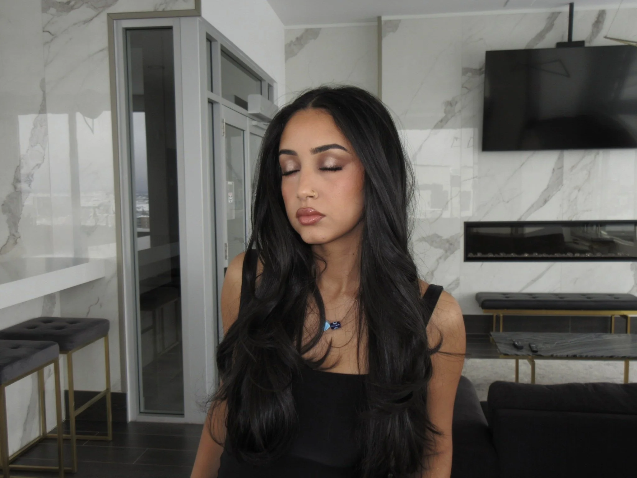 A woman with long dark hair and a nose ring has her eyes closed to show her full glam makeup. She is in a room with modern decor, marble walls, and a mounted television.