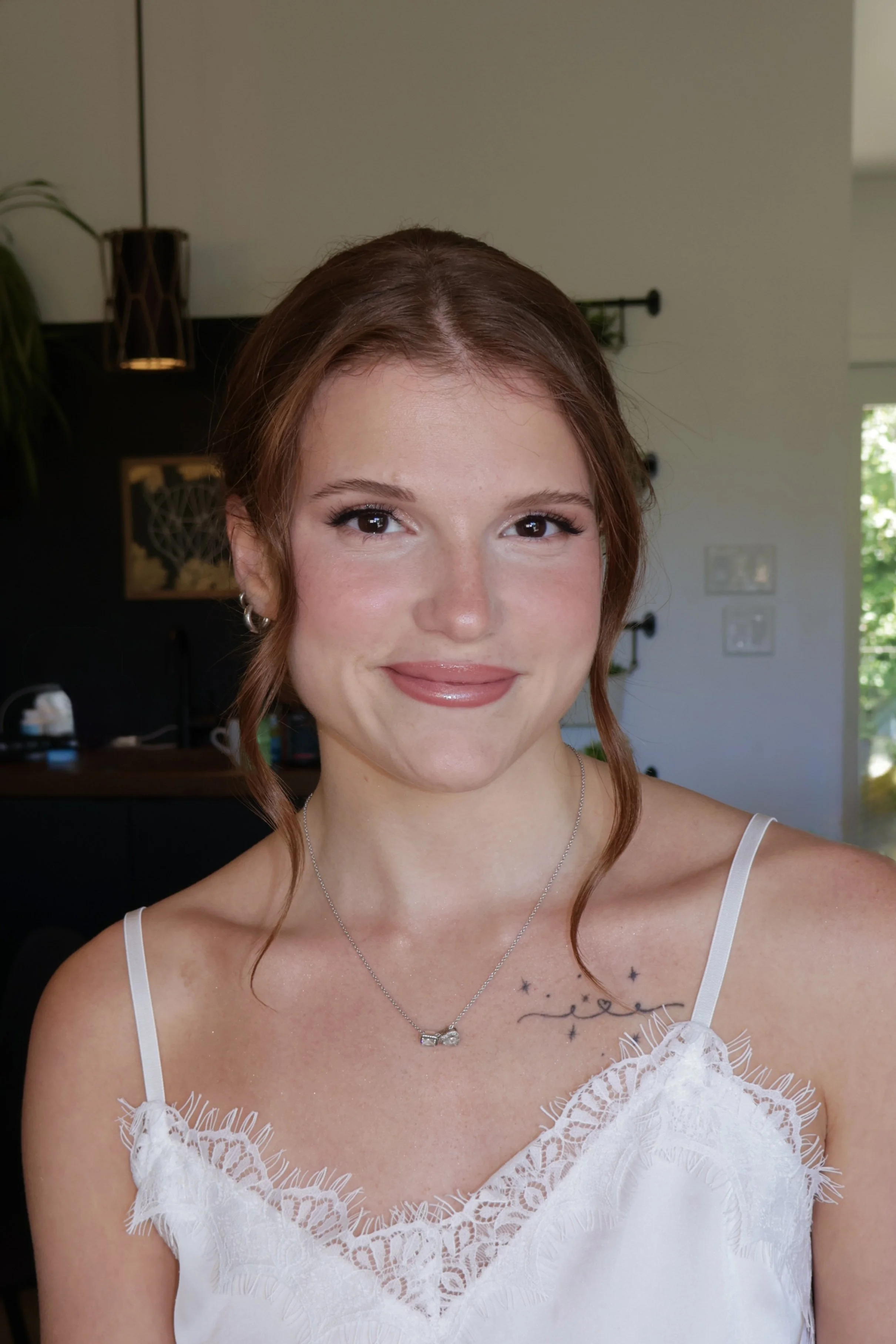 Close-up of a young woman with auburn hair, wearing a white lace dress with spaghetti straps, and a silver necklace. She is smiling softly to show off her bridal makeup, with a background featuring indoor decor and a window with greenery outside.