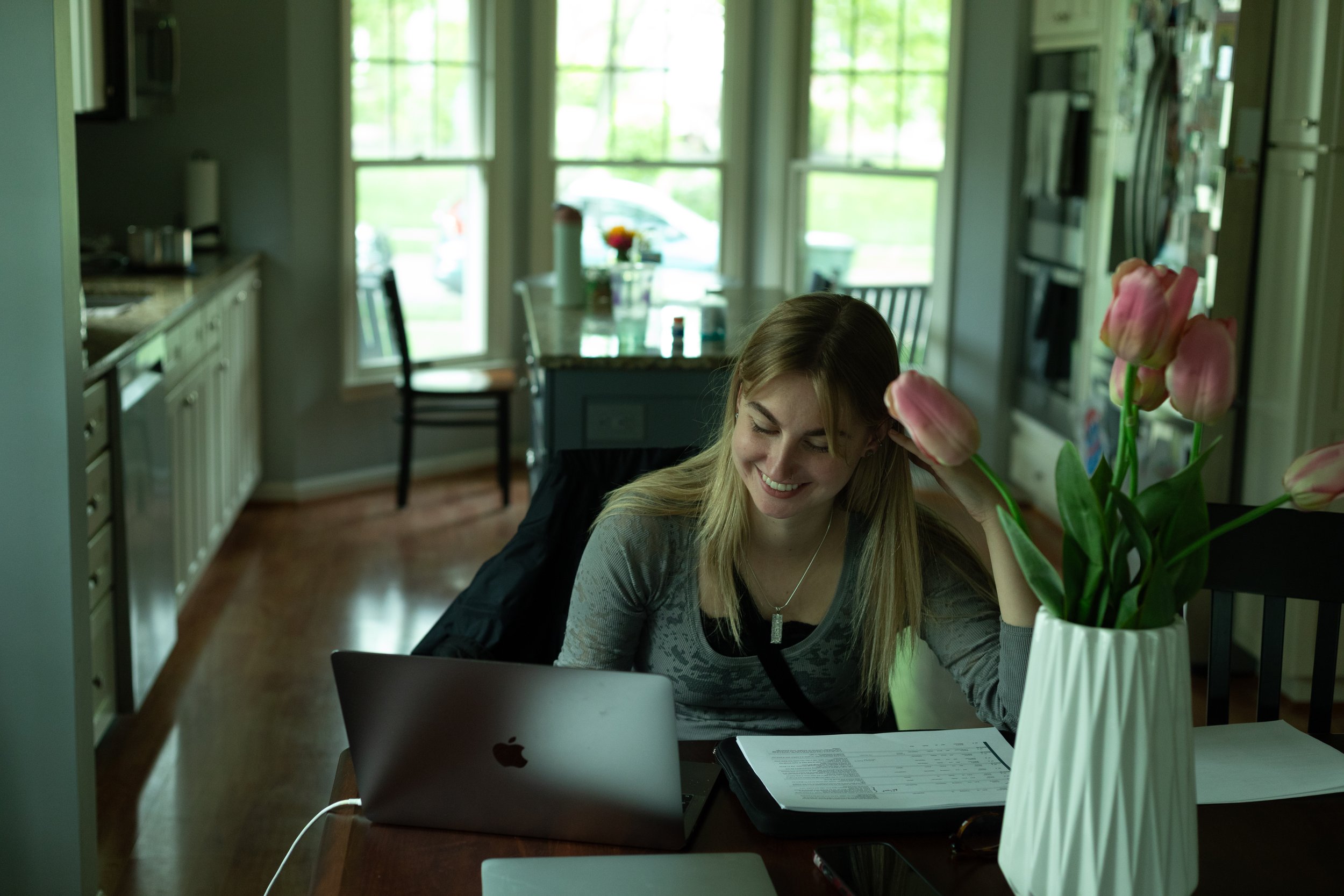 A woman with long blonde hair smiling while working on a laptop at a dining table in a sunlit kitchen, with a large flower vase containing pink tulips in the foreground.