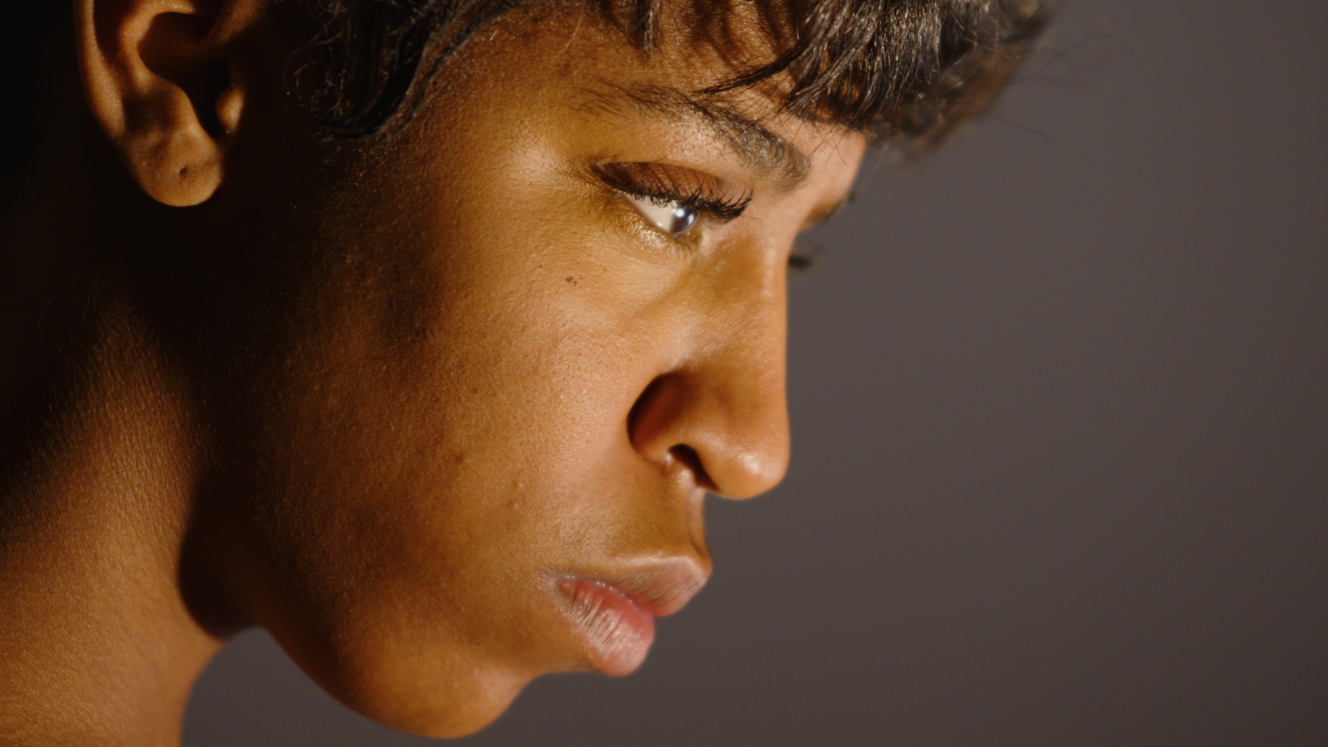 Close-up side profile of a young woman with short dark hair, smooth brown skin, and closed lips, against a neutral background.