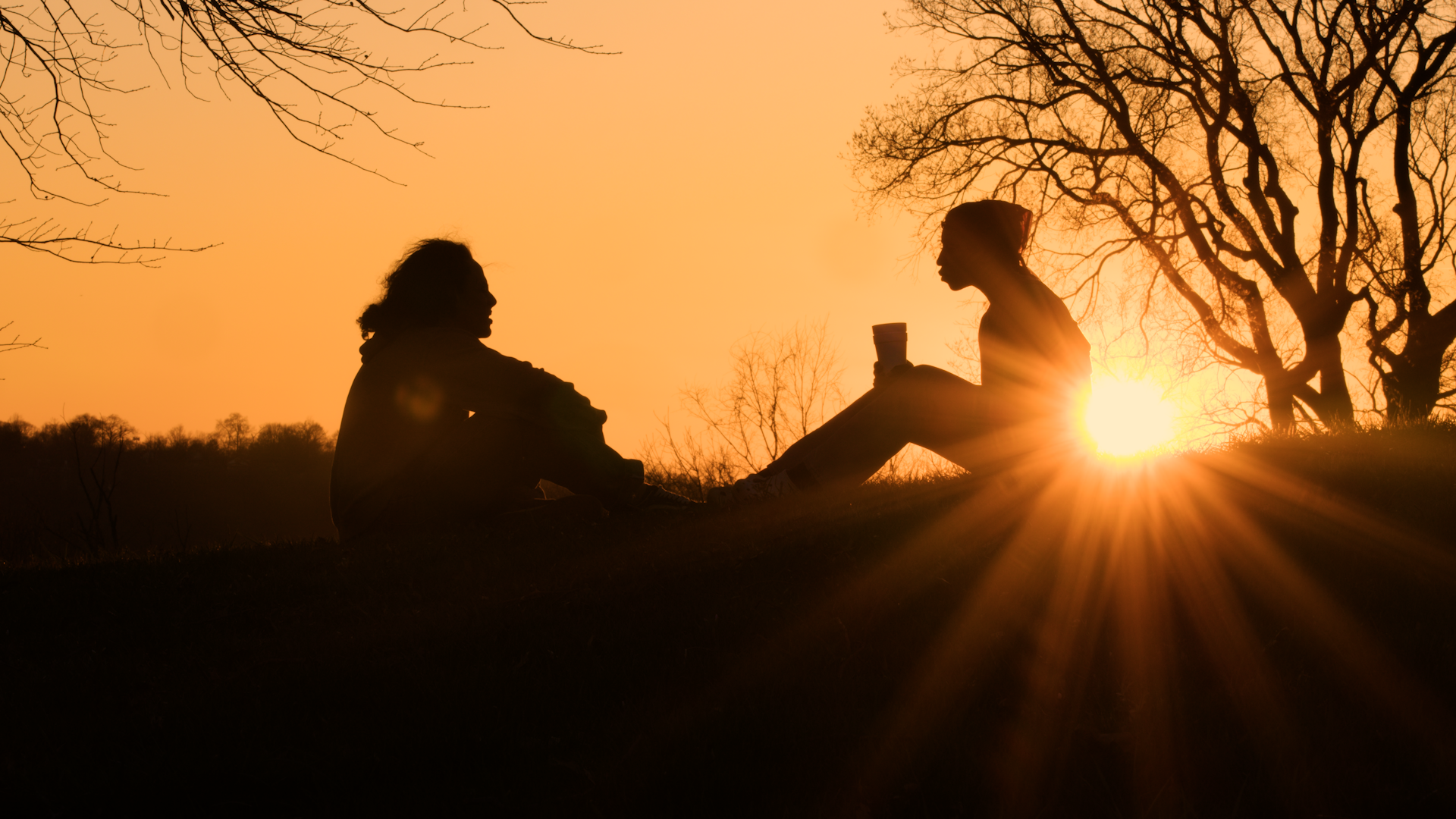 Silhouettes of two women sitting on the ground outdoors during sunset, with leafless trees and a bright orange sky in the background.
