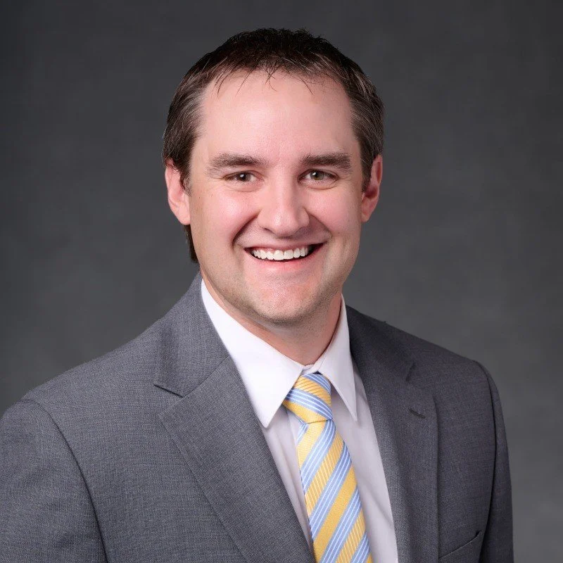 Smiling man in a gray suit and striped yellow-and-blue tie against a dark gray background