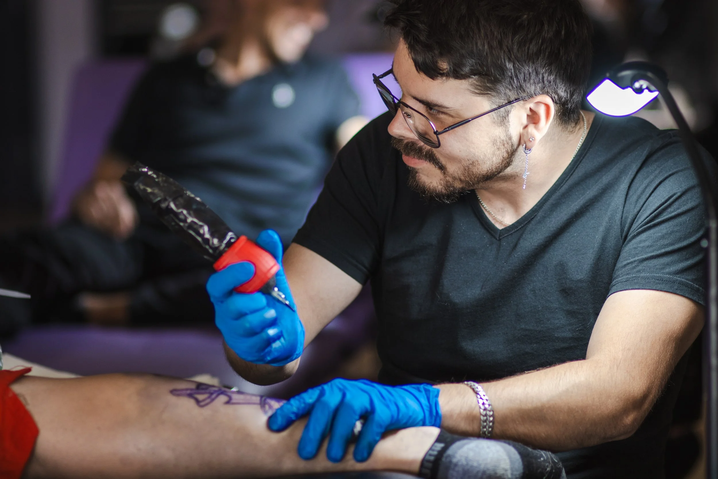 Tattoo artist wearing blue gloves tattoos a client’s forearm with a tattoo machine.