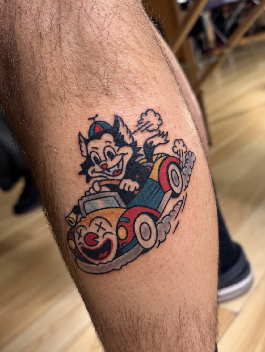 Colorful tattoo of a cartoon mouse driving a small red-and-blue race car on a person’s leg. Work by Ruben.