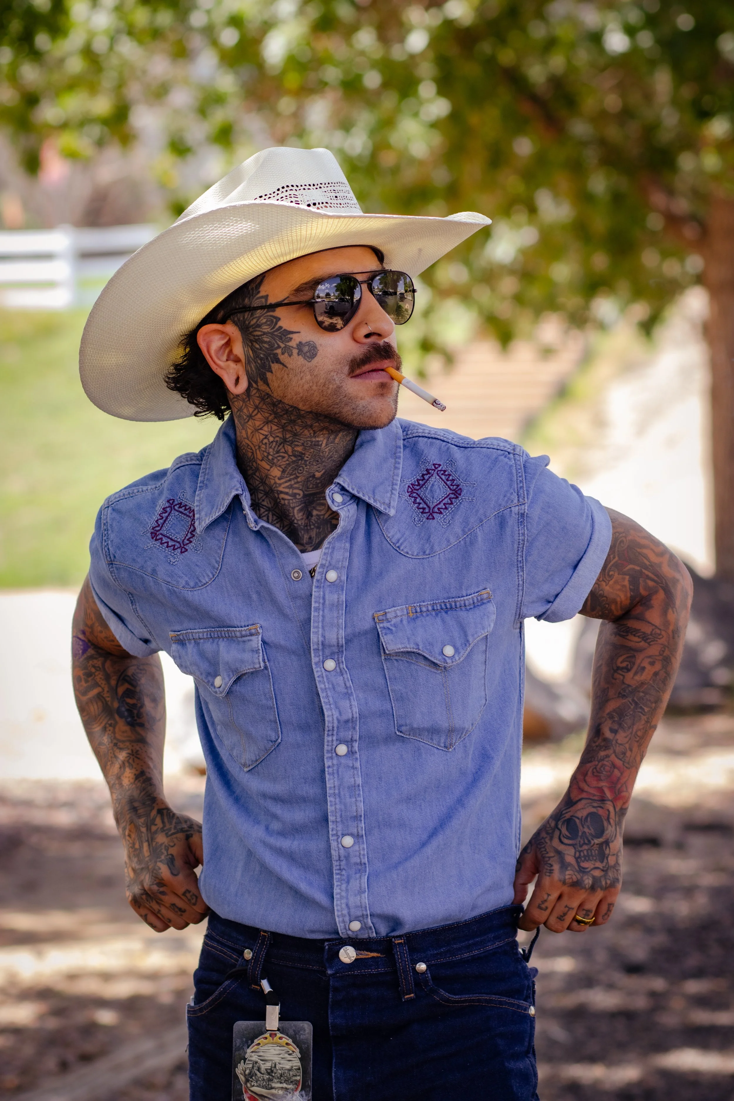 Tattooed man (Ruben) wearing a white cowboy hat, sunglasses, and blue denim shirt while smoking outdoors