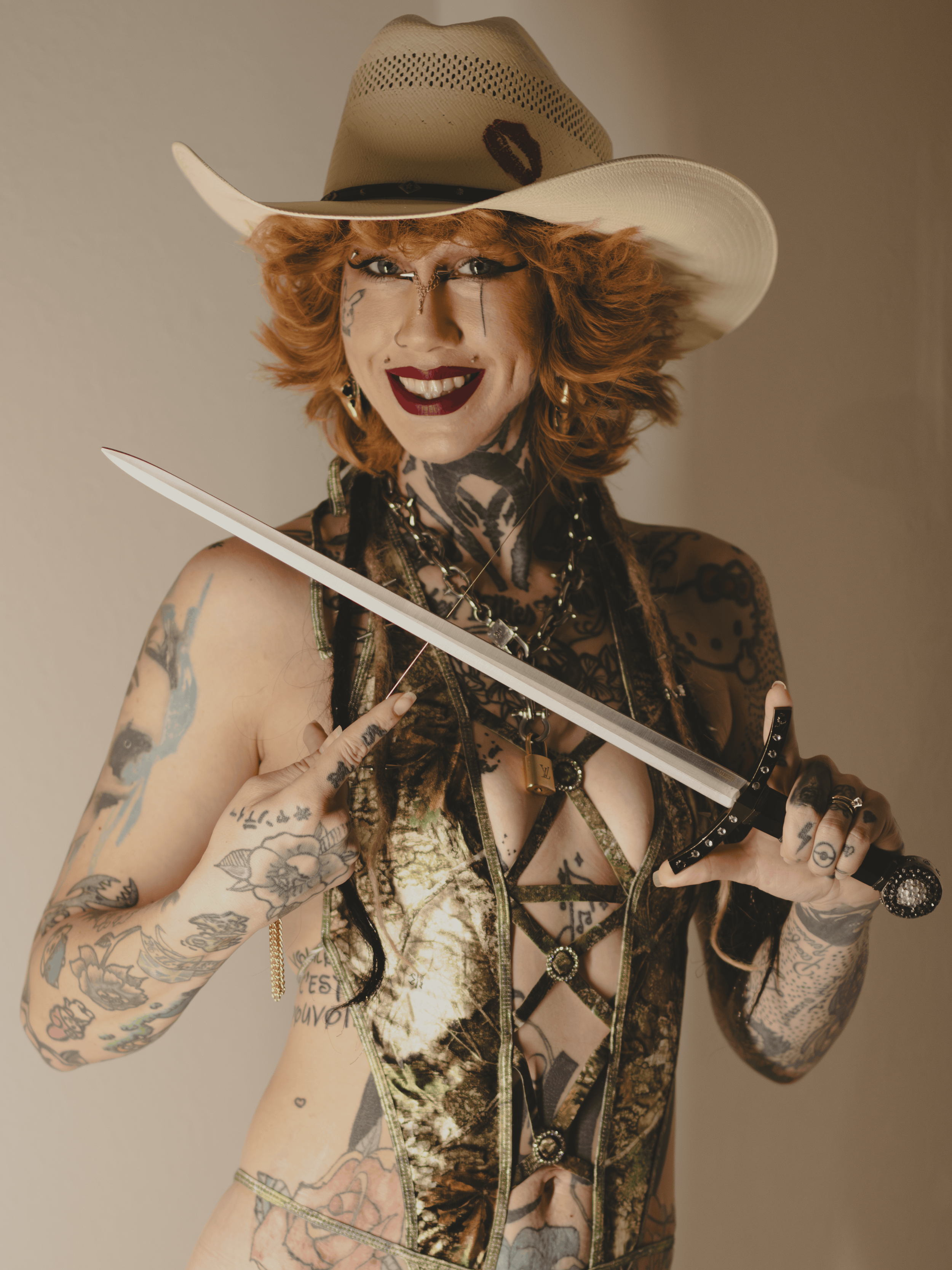 Smiling tattooed woman (Hannah) with reddish hair wearing a white cowboy hat and holding a sword