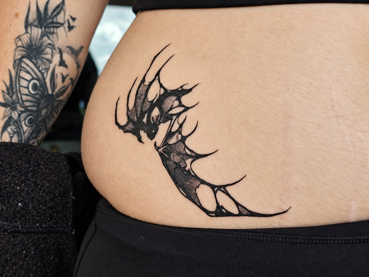 Close-up of a person’s hip showing a black ink abstract bat tattoo, with a floral and bat tattoo visible on the arm in the background.
